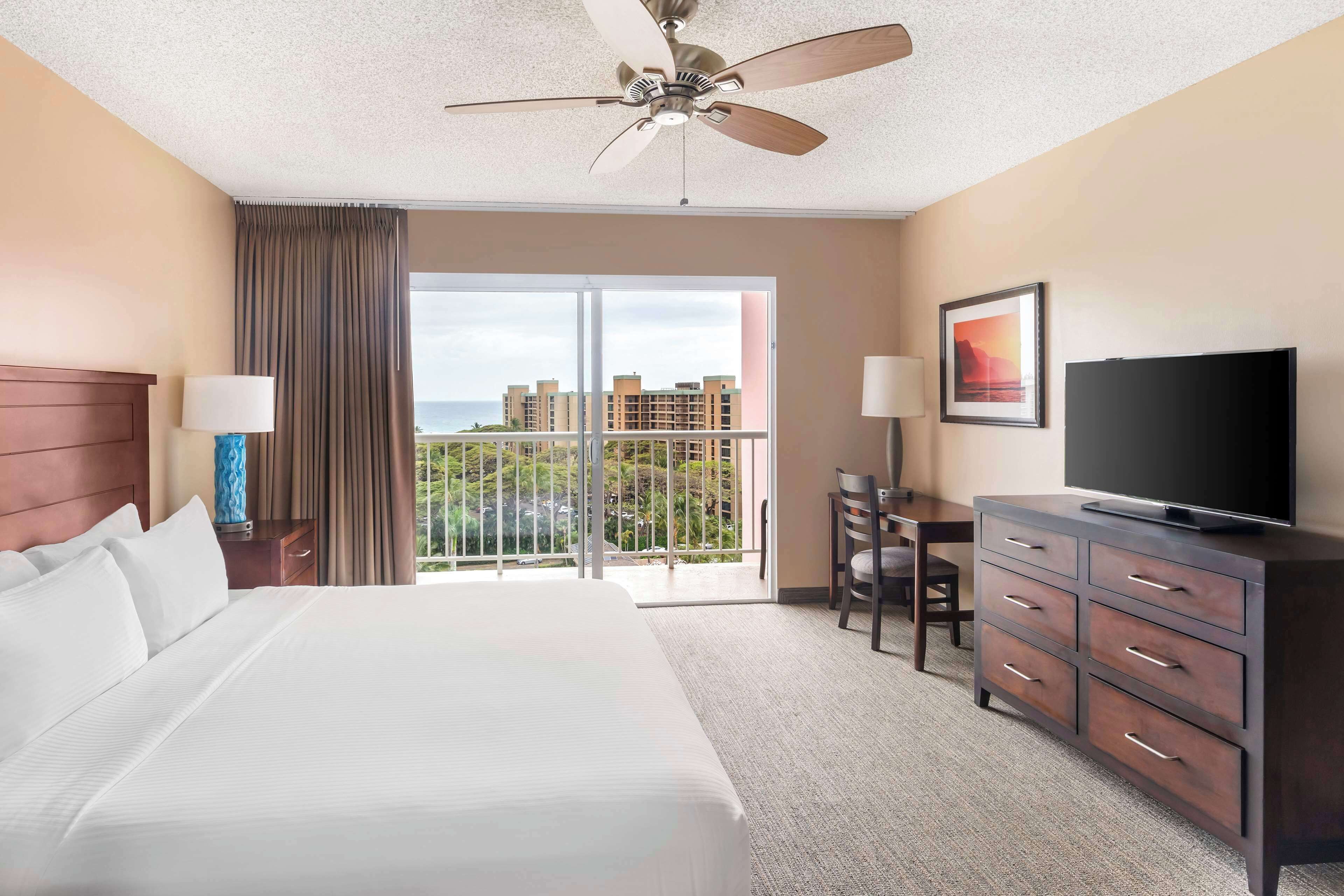 A hotel room with a large bed, a ceiling fan, a television on a dresser, a desk with a chair, and a view of buildings through the window.