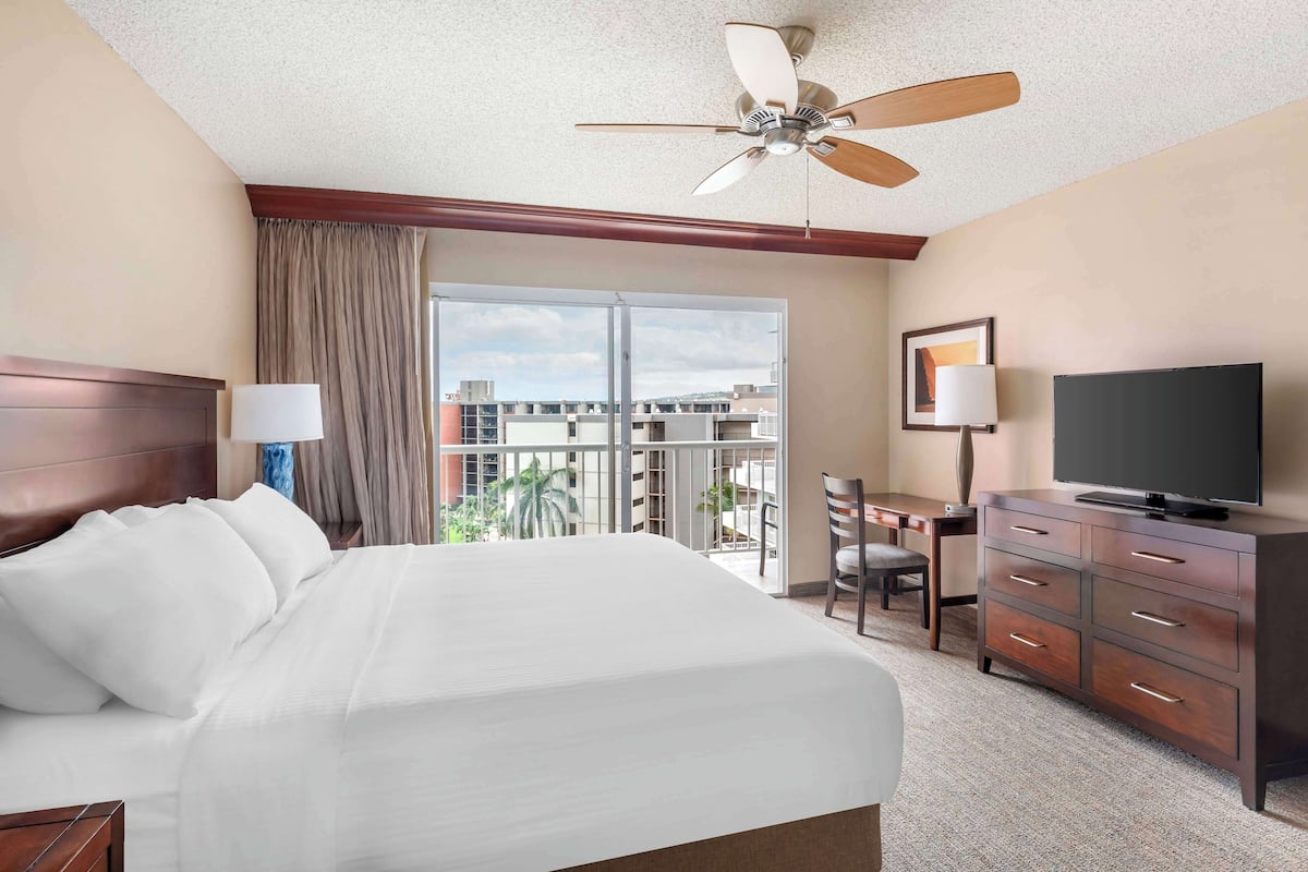 A hotel room with a large bed, a desk with a chair, a television, and a balcony with a view of buildings.