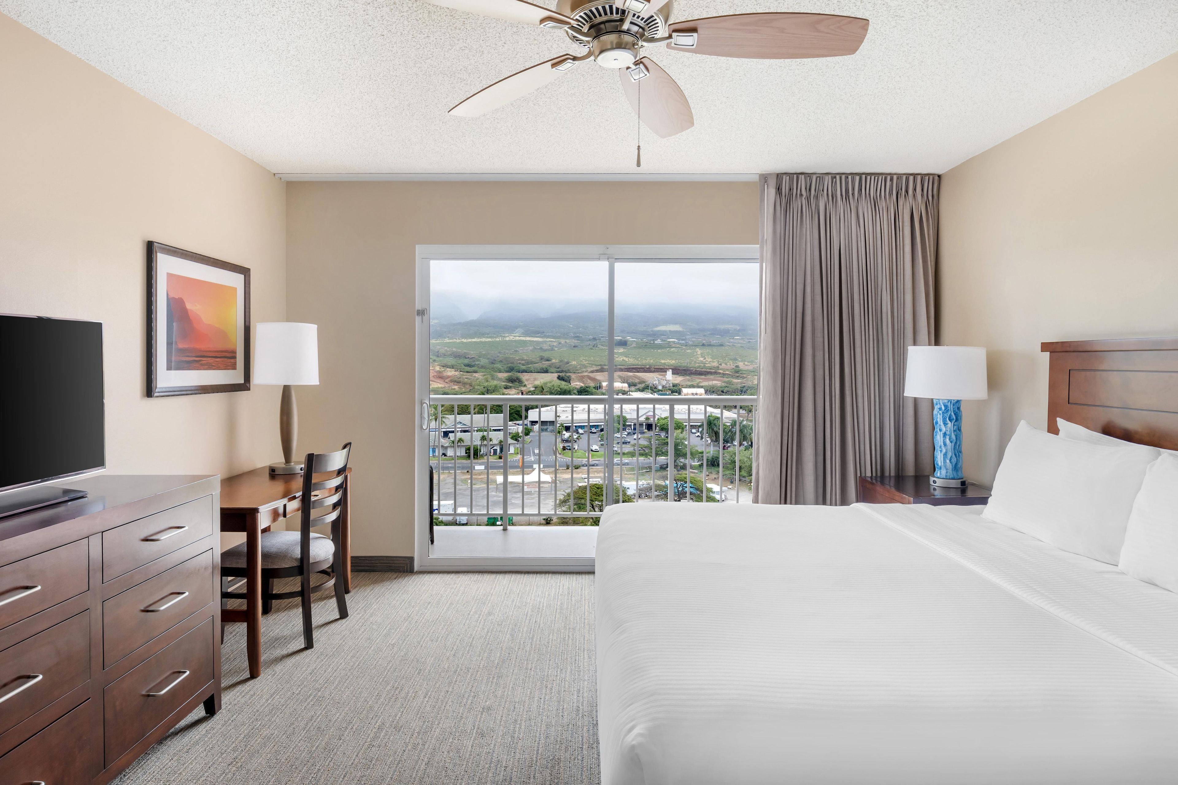 A hotel room with a bed, a desk with a chair, a television, and a view of a cityscape through a large window.