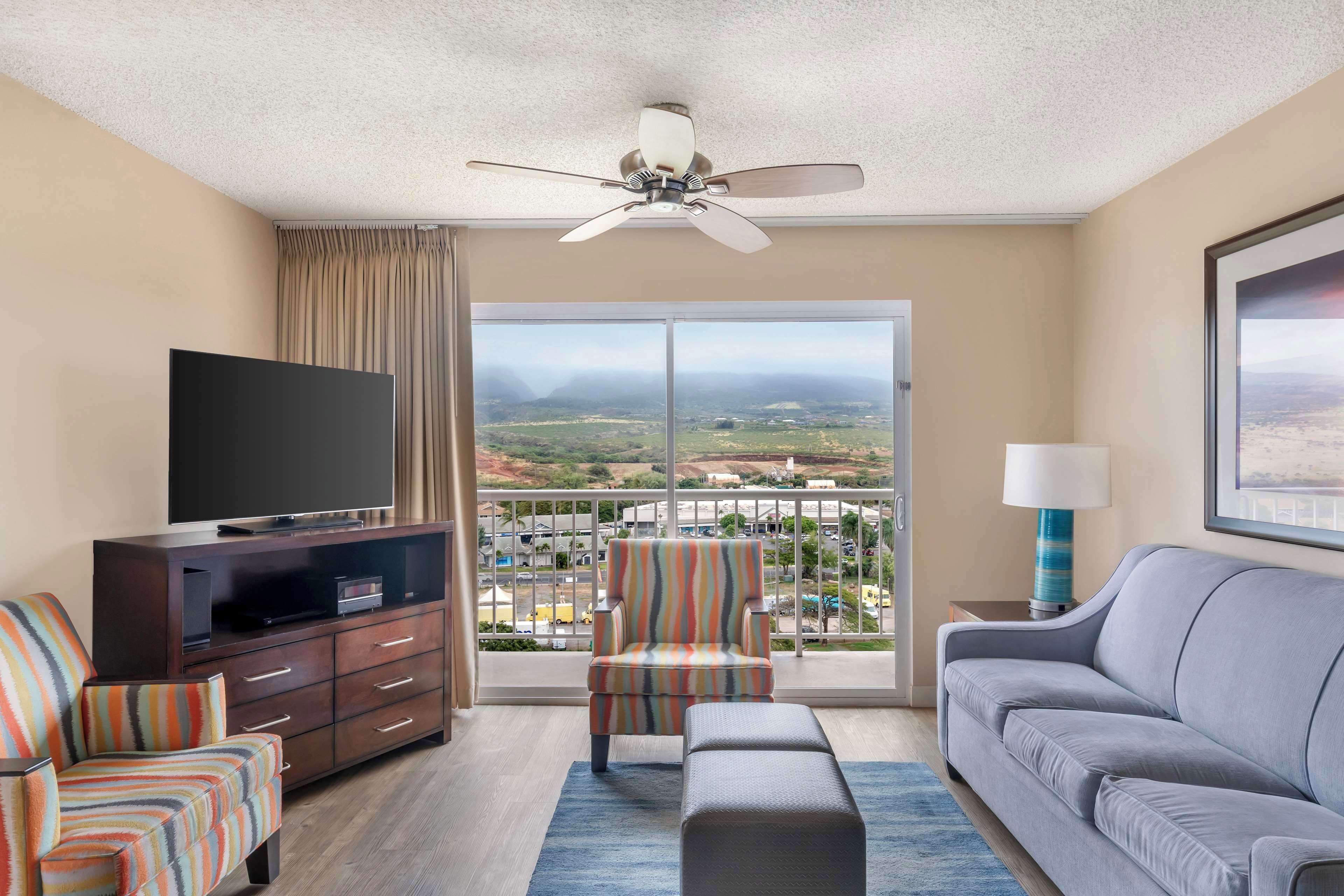 A hotel room with a large window, a ceiling fan, a flat-screen TV, a sofa, a chair, and a small table with a lamp.