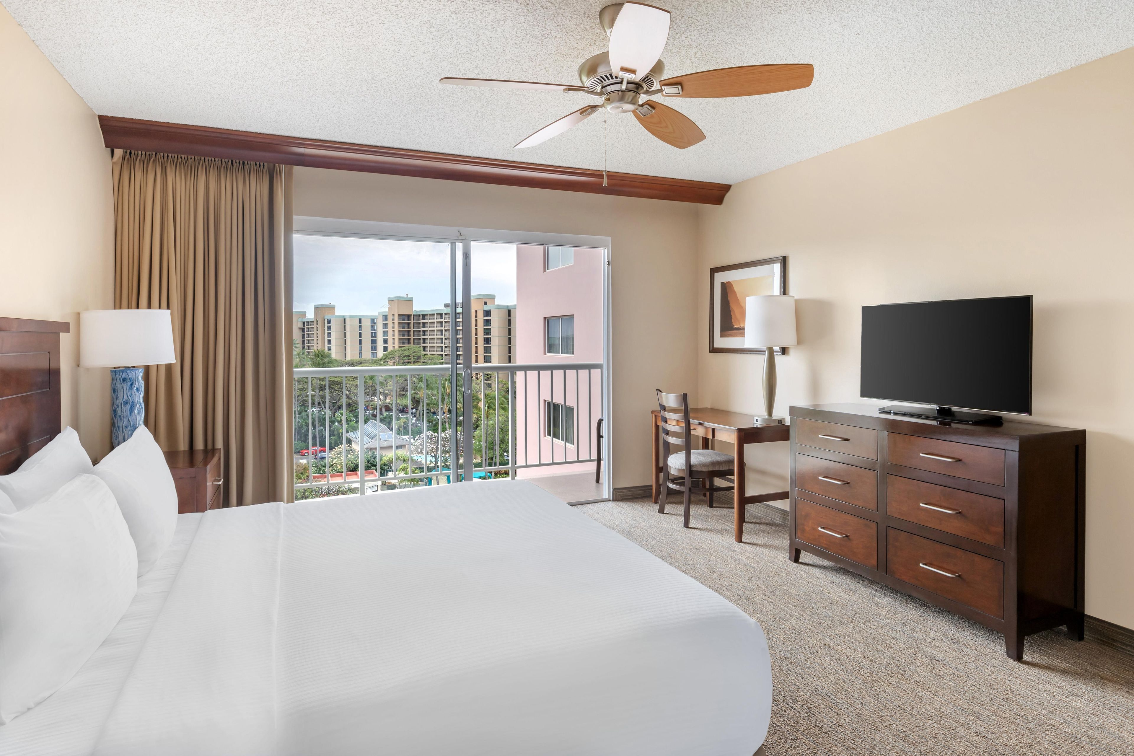 A hotel room with a large bed, a television, a desk, and a balcony with a view of buildings.