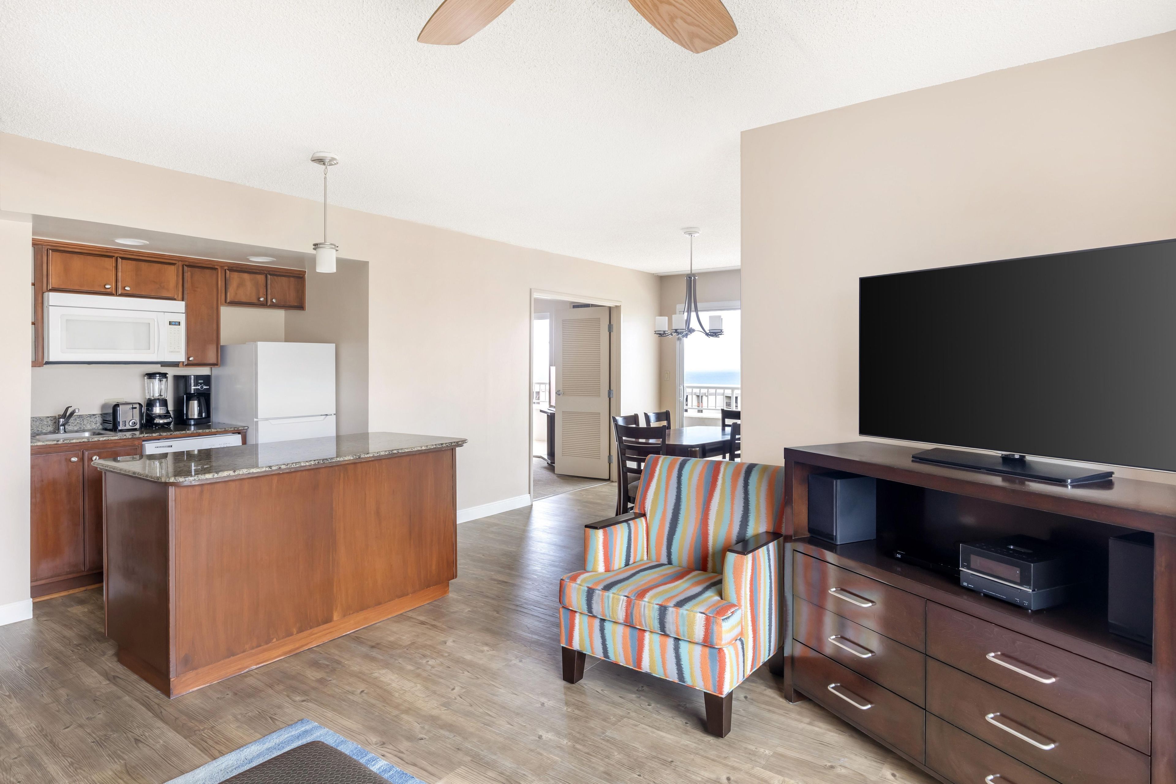 A modern kitchen with wooden cabinets, a large island, and a dining area with a colorful striped chair.