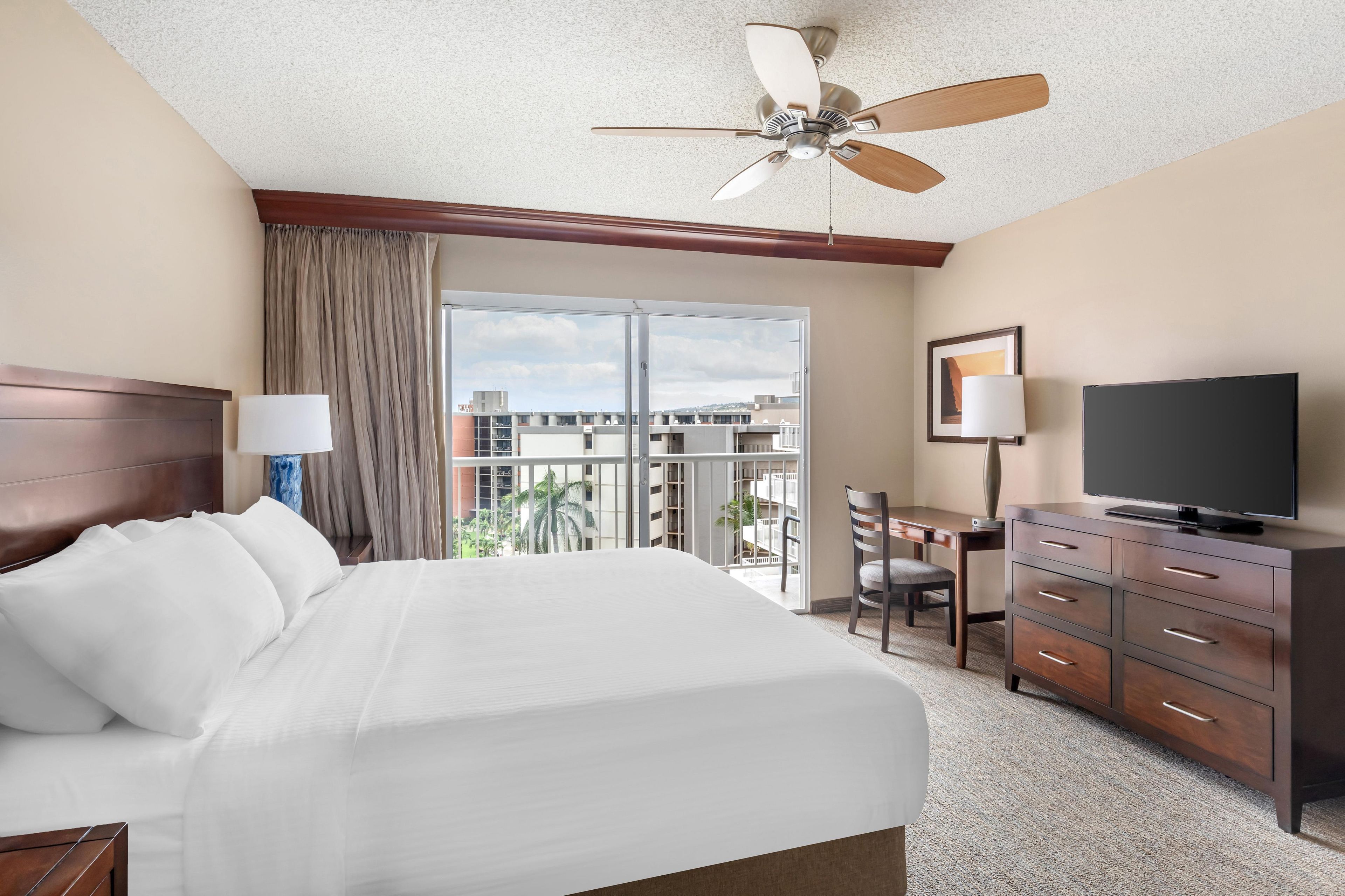 Habitación de hotel con cama, escritorio, televisor, ventilador de techo y vista a la ciudad.