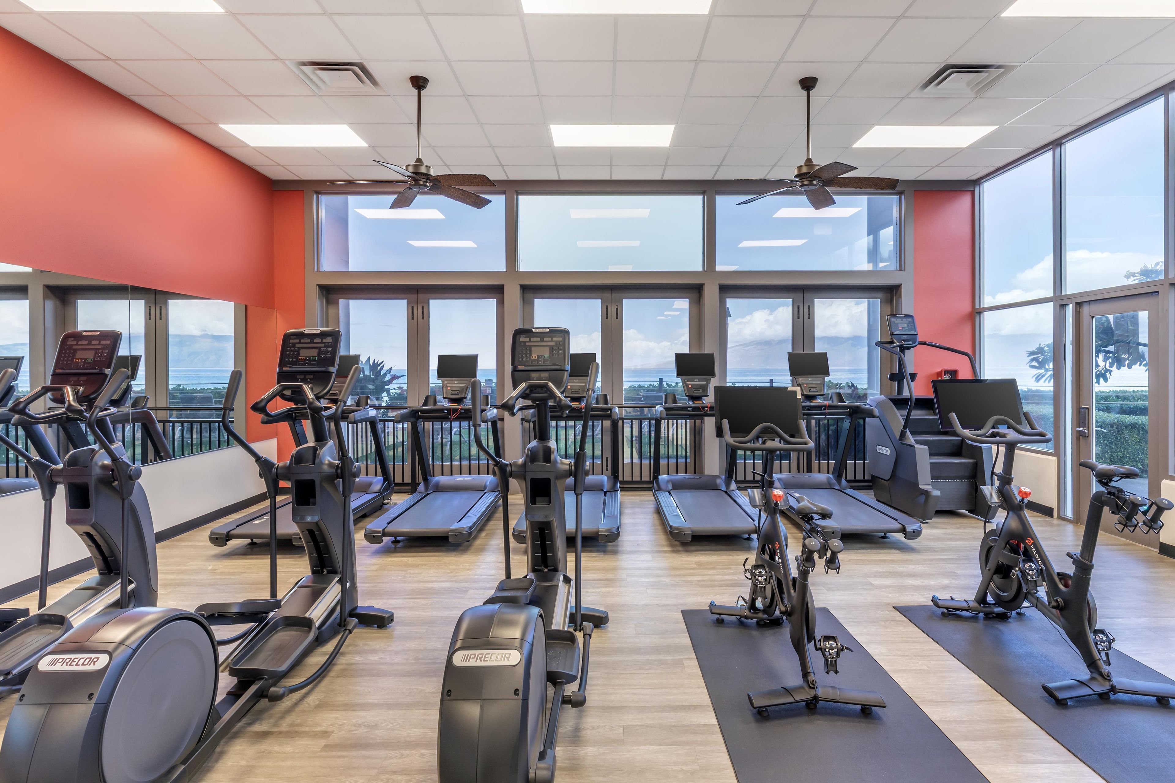 A gym with multiple exercise machines, large windows, and ceiling fans.