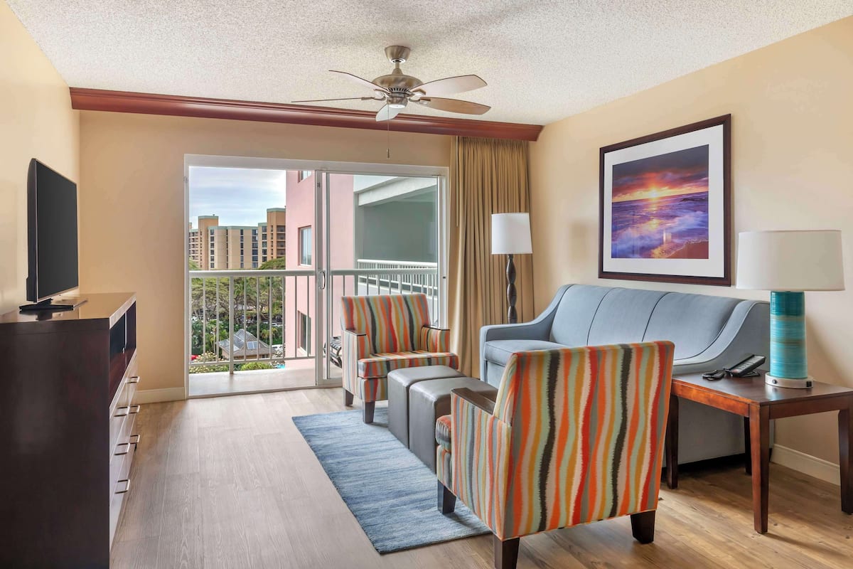 A hotel room with a balcony, a sofa, striped armchairs, a TV, and a painting.