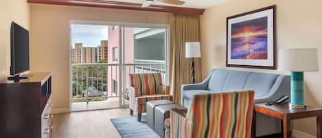 A hotel room with a balcony, a sofa, striped armchairs, a TV, and a painting.