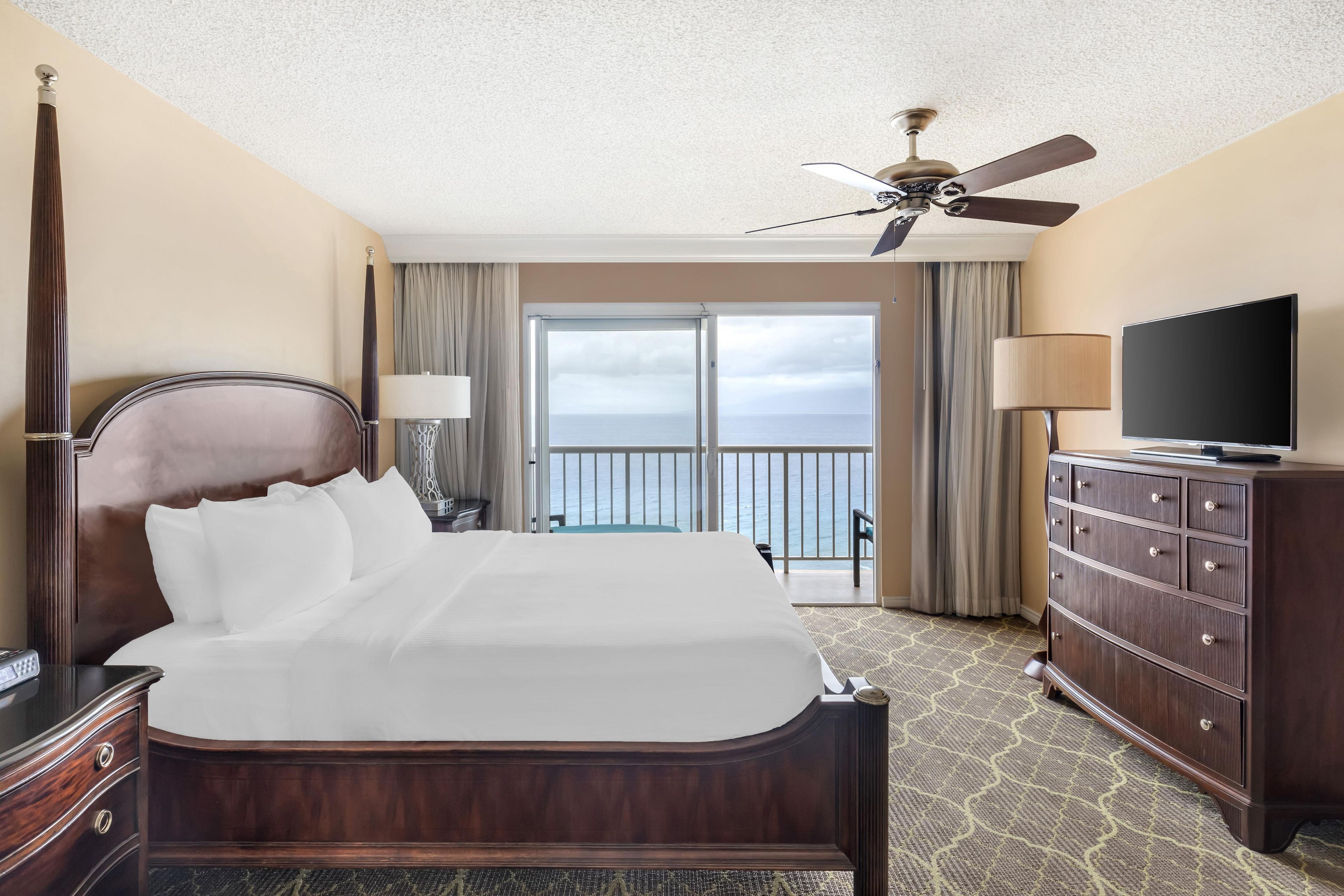 A hotel room with a large bed, a television, a dresser, and a balcony with a view of the sea.