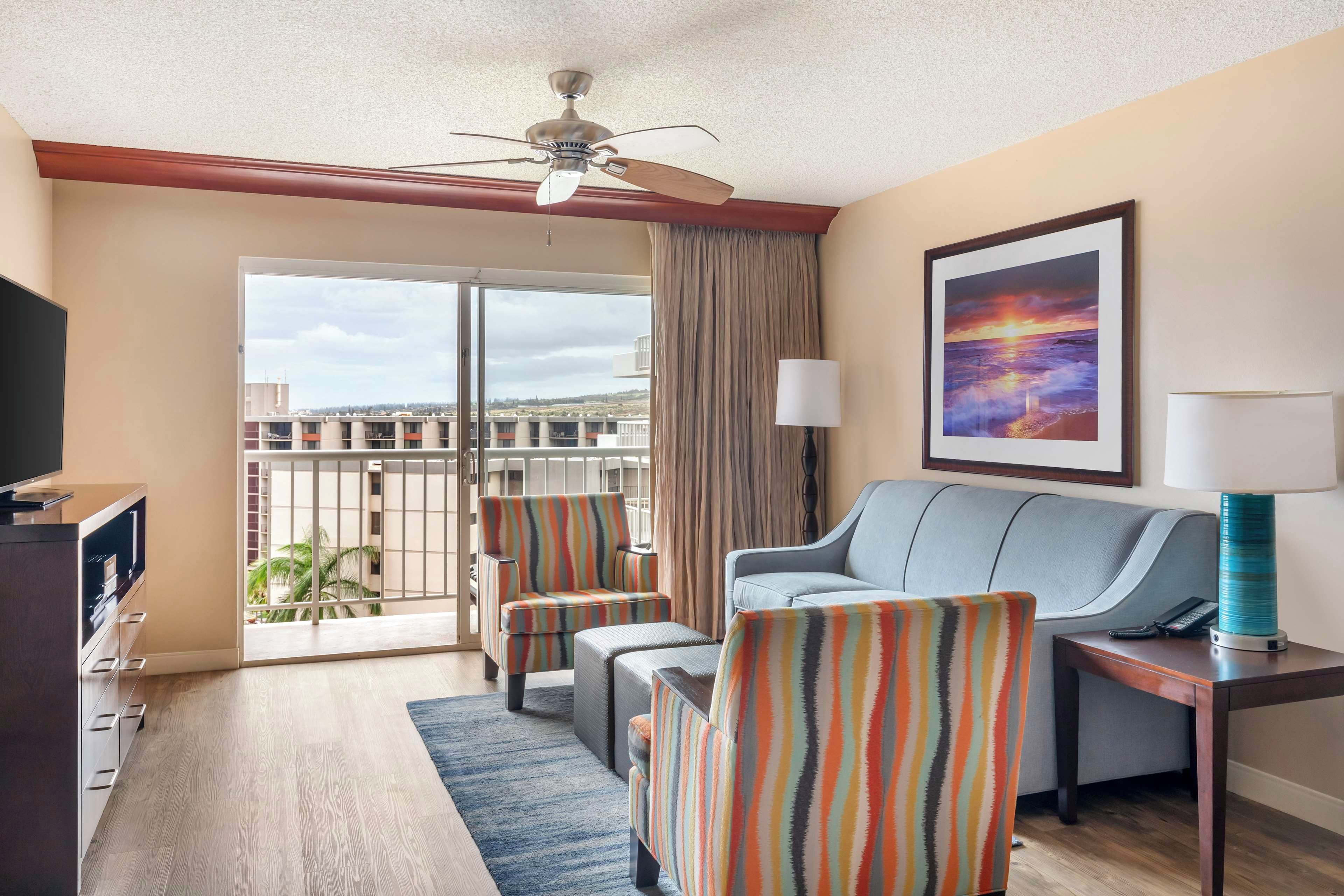 A hotel room with a balcony, a sofa, two striped armchairs, a TV, and a painting of a sunset.
