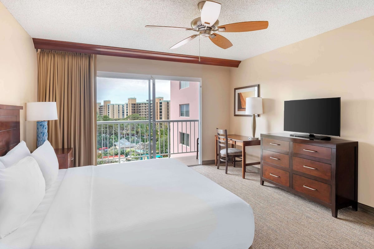 A hotel room with a large bed, a desk with a chair, a television, and a balcony with a view of buildings.