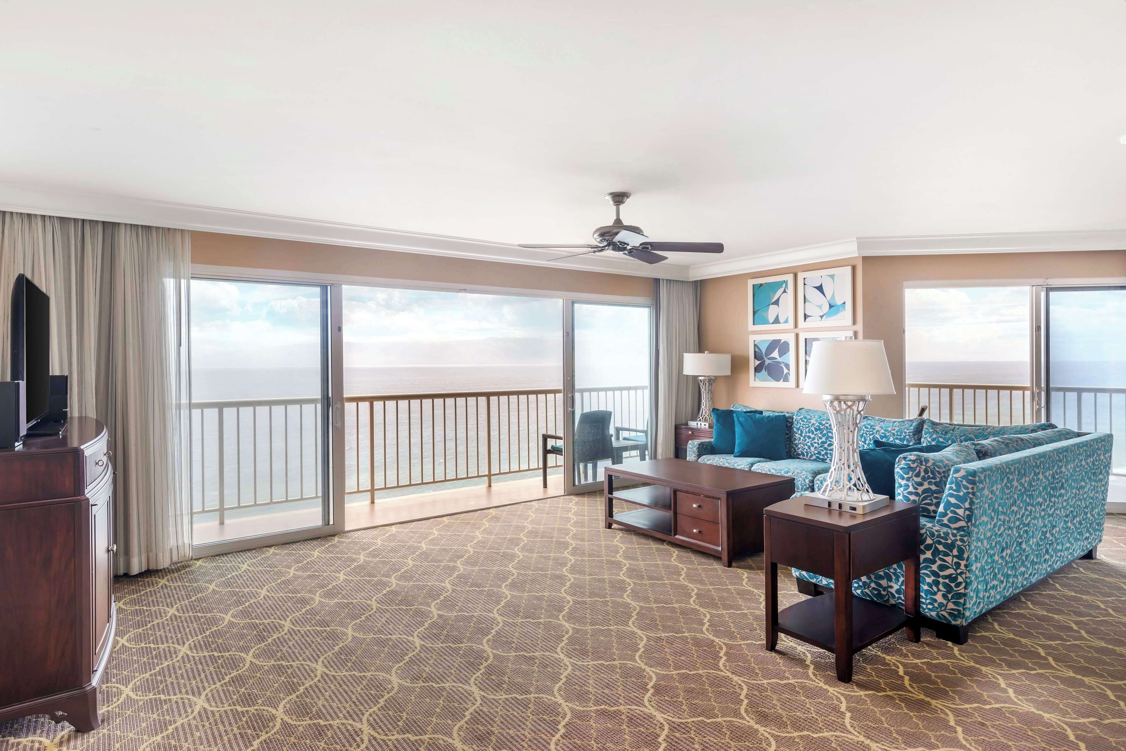 A hotel room with a large balcony, a sofa, a coffee table, a TV, and a ceiling fan.