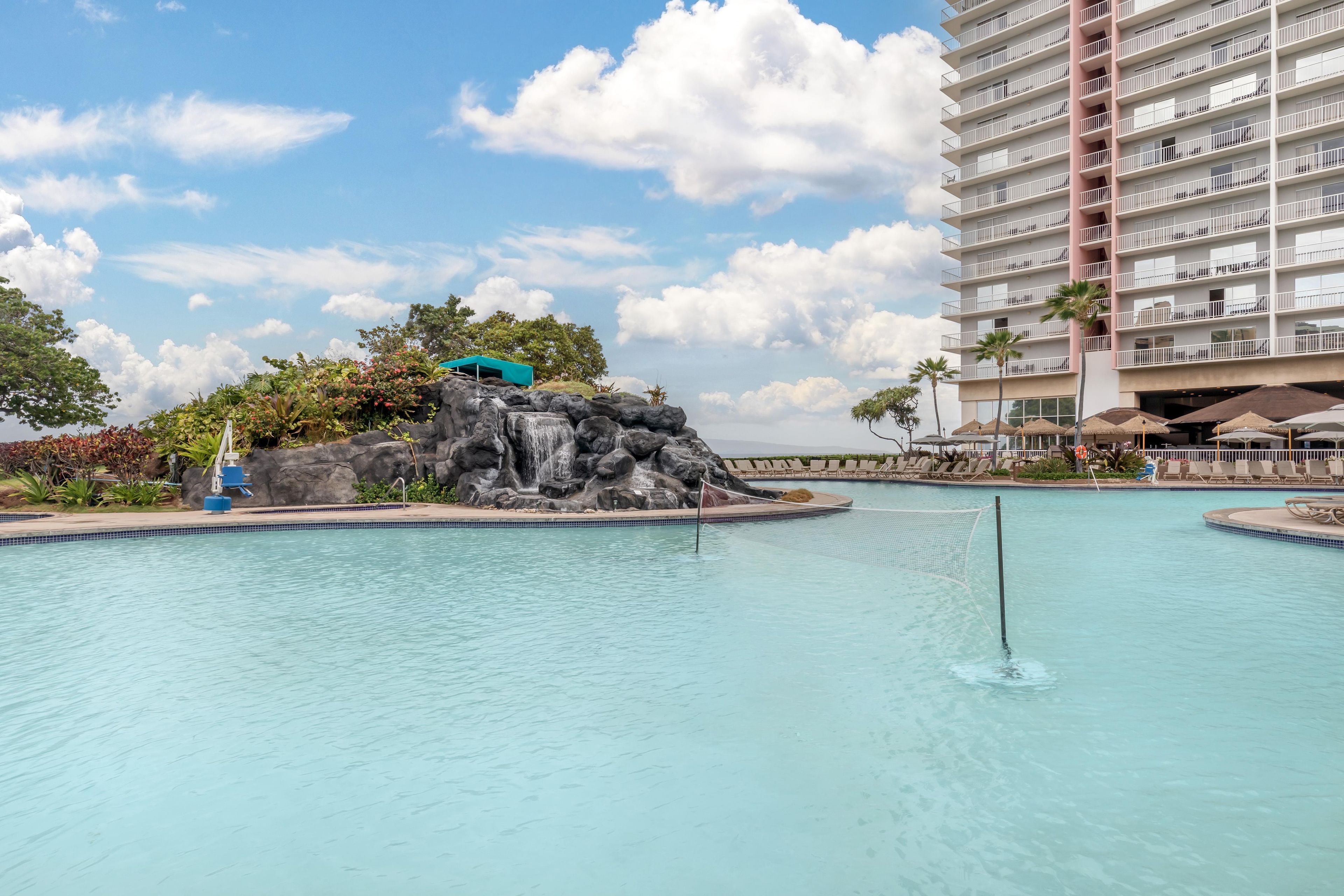 Una piscina con caída de agua, rodeada de sillas de descanso y un edificio de varios pisos.