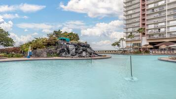 A swimming pool with a waterfall feature, surrounded by lounge chairs and a multi-story building.