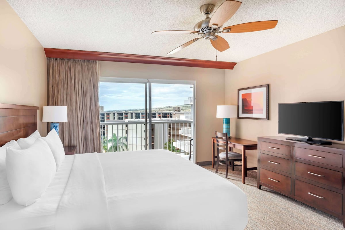 A hotel room with a large bed, a desk with a chair, a television, and a balcony with a view of the city.