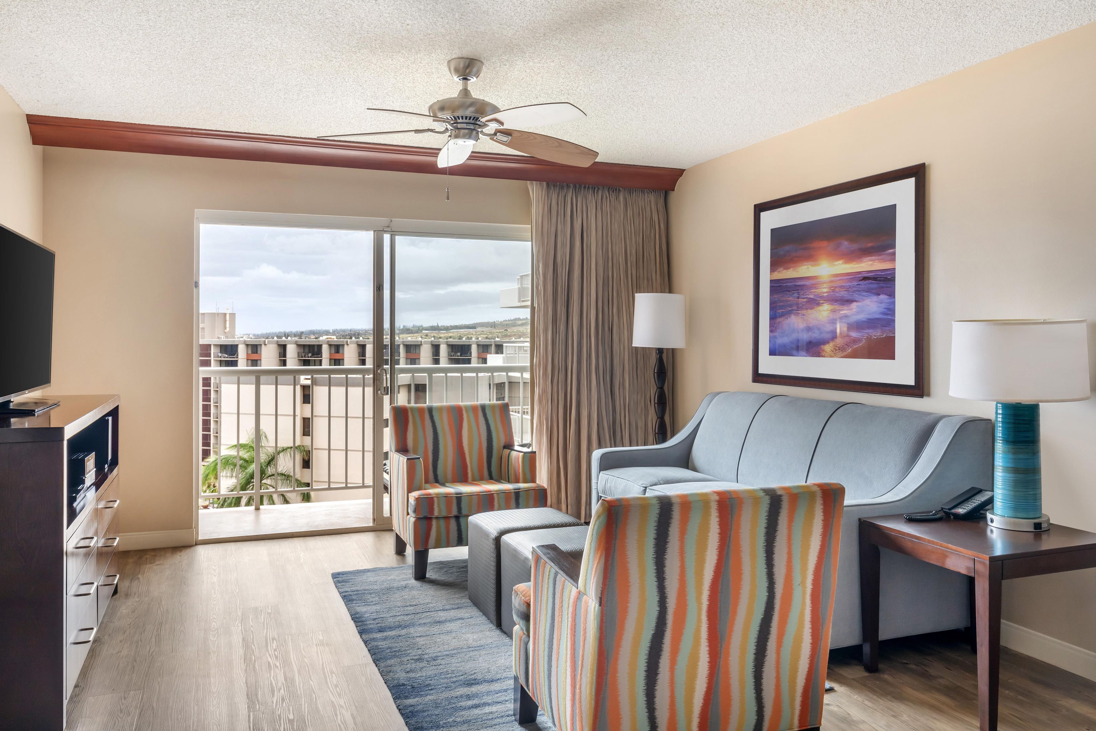 Habitación de hotel con balcón, sofá, dos sillones, televisor, lámpara y un cuadro.