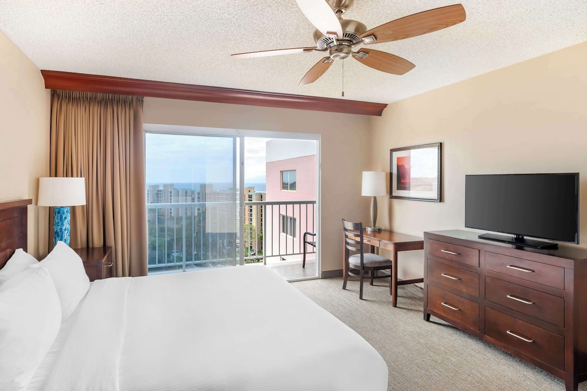 A hotel room with a large bed, a desk with a chair, a television, and a balcony with a city view.