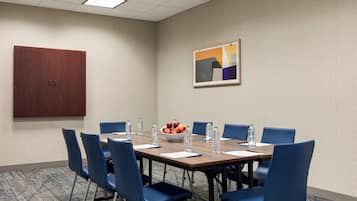 A conference room with a wooden table, blue chairs, a fruit bowl, water bottles, and a framed abstract painting on the wall.
