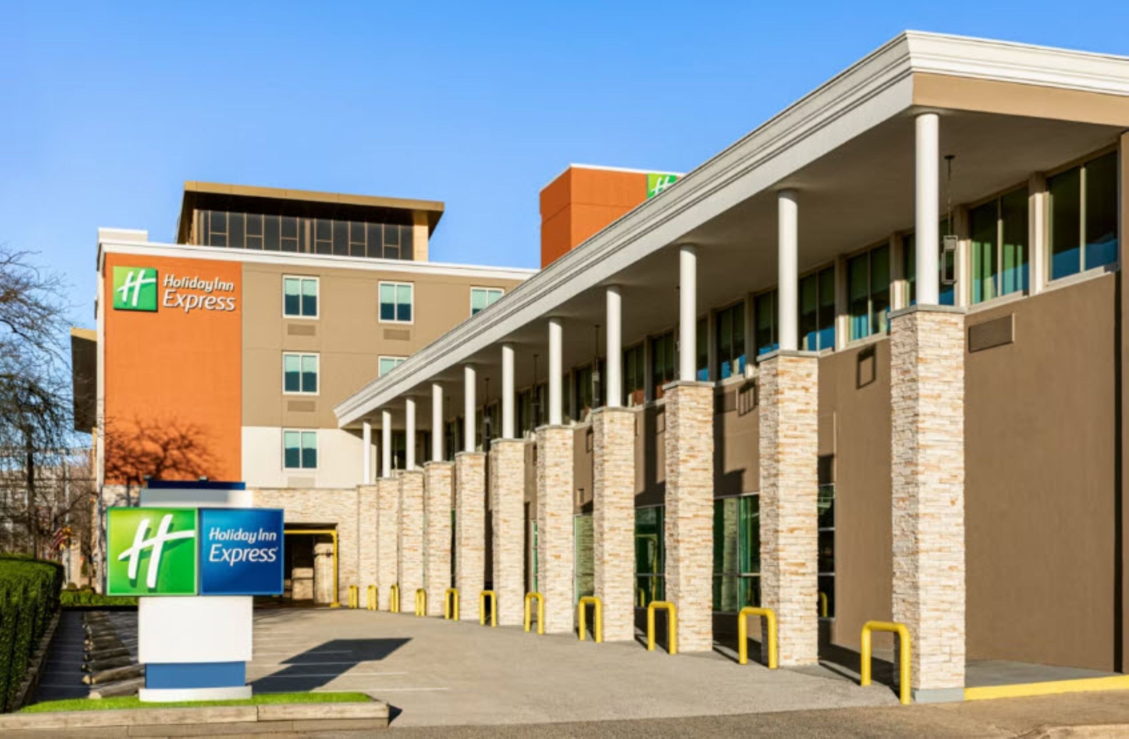 A Holiday Inn Express hotel with a modern design, featuring a central atrium with columns, a parking lot, and a clear sky.