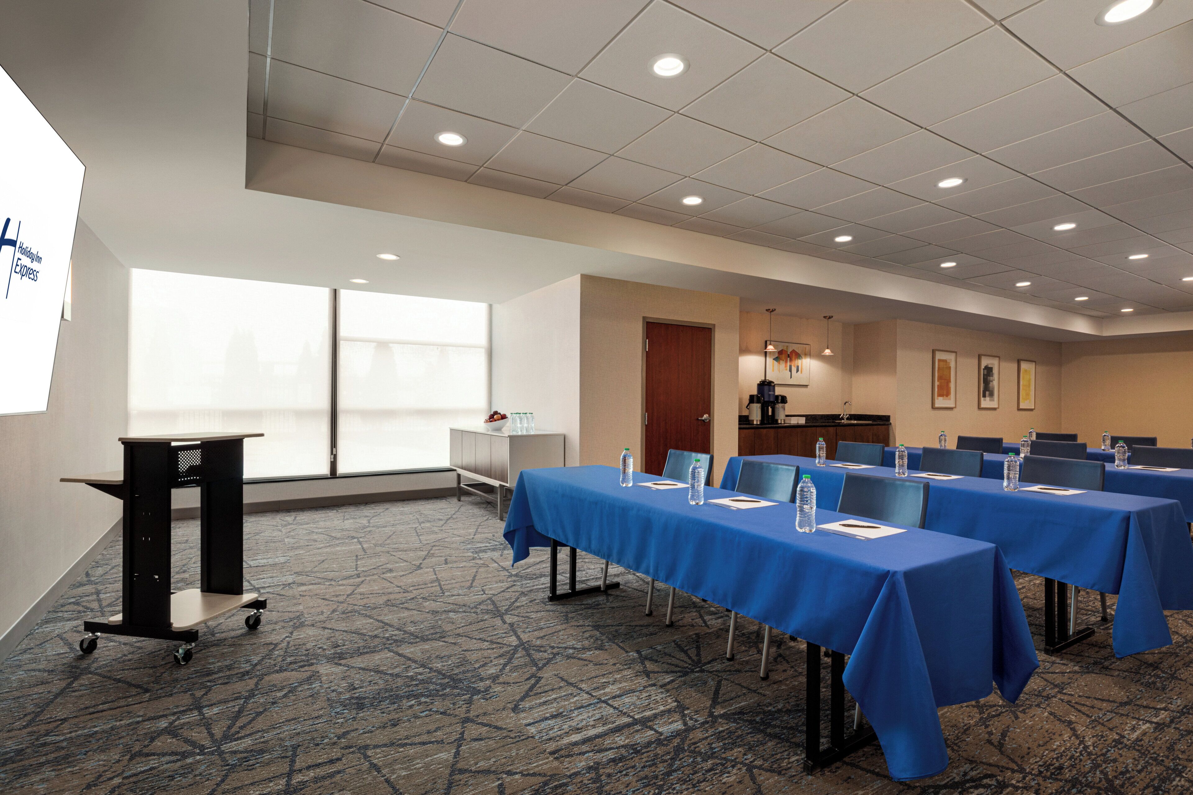 A conference room with a long table covered in blue, equipped with water bottles and notepads, and a projector screen on the left.