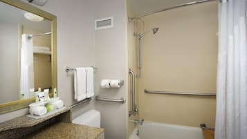 Un baño con bañera, inodoro, lavamanos con artículos de aseo, espejo y ducha con barras de apoyo.