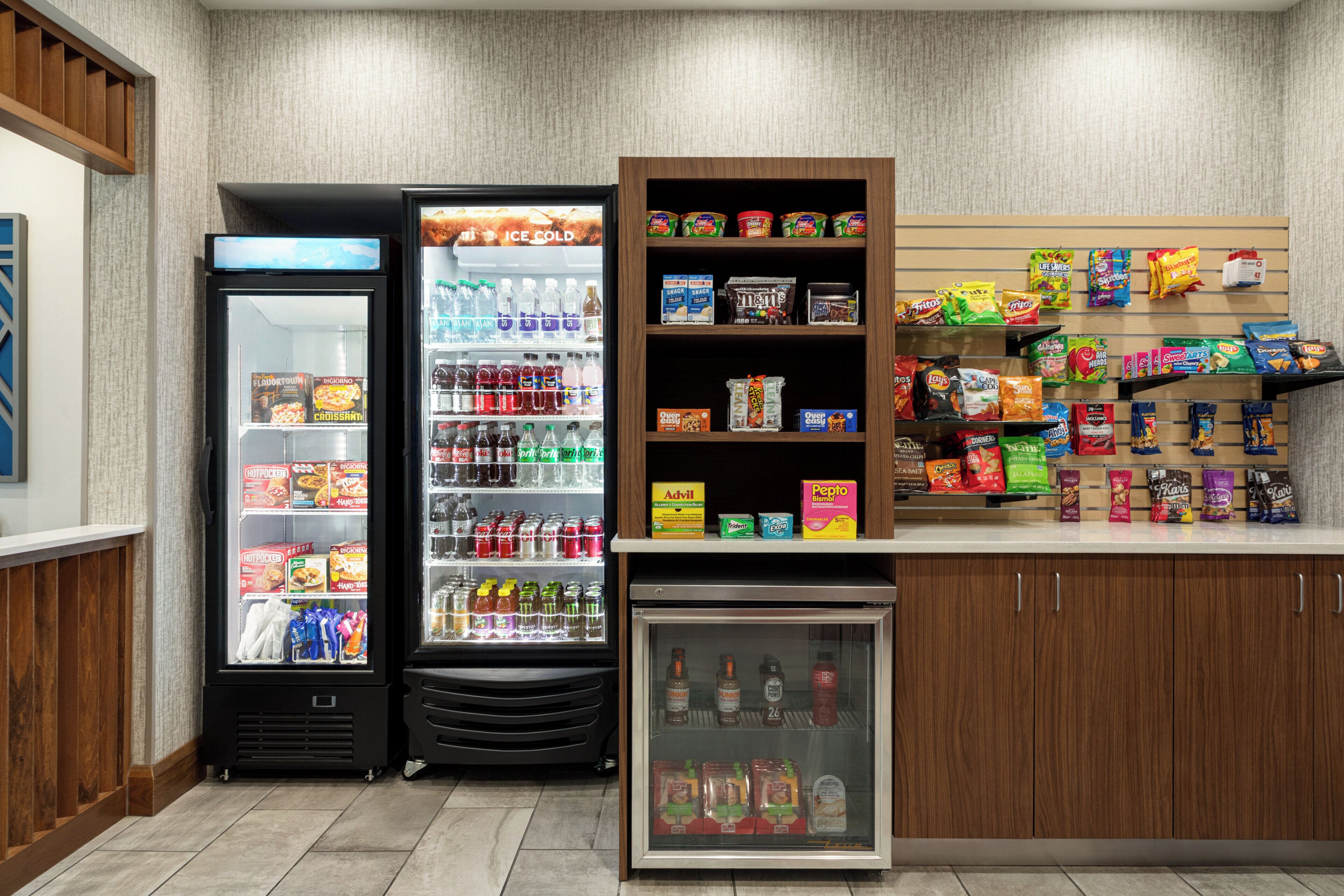 A self-service beverage and snack area with refrigerated drink dispensers, a small refrigerator, and shelves stocked with various snacks and beverages.