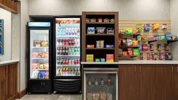 A self-service beverage and snack area with refrigerated drink dispensers, a small refrigerator, and shelves stocked with various snacks and beverages.