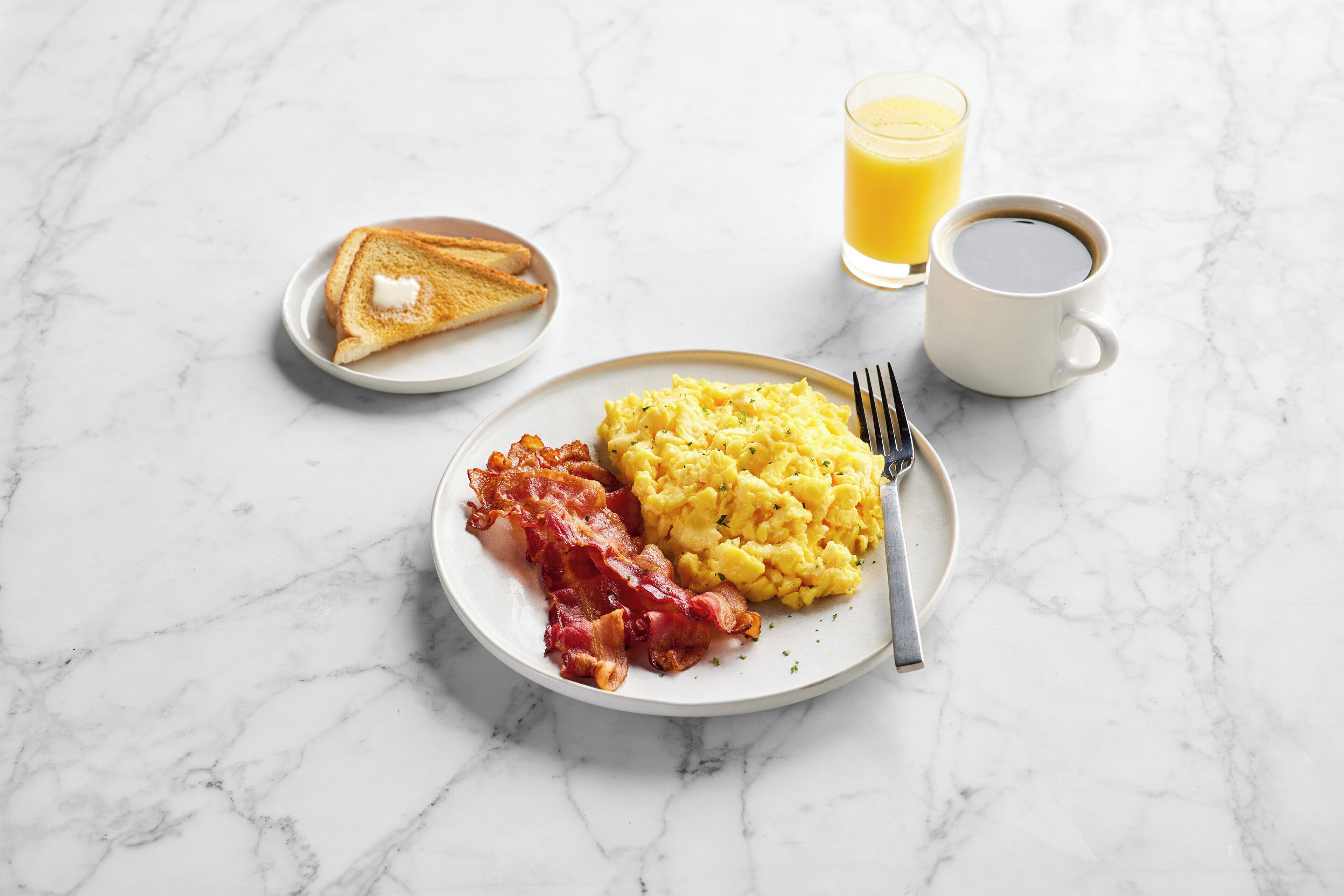 Une assiette de petit-déjeuner avec des œufs brouillés, du bacon, des toasts, un verre de jus d’orange et une tasse de café, le tout posé sur une surface en marbre.