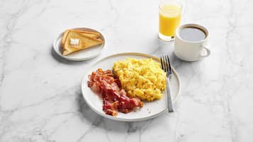 Une assiette de petit-déjeuner avec des œufs brouillés, du bacon, des toasts, un verre de jus d’orange et une tasse de café, le tout posé sur une surface en marbre.