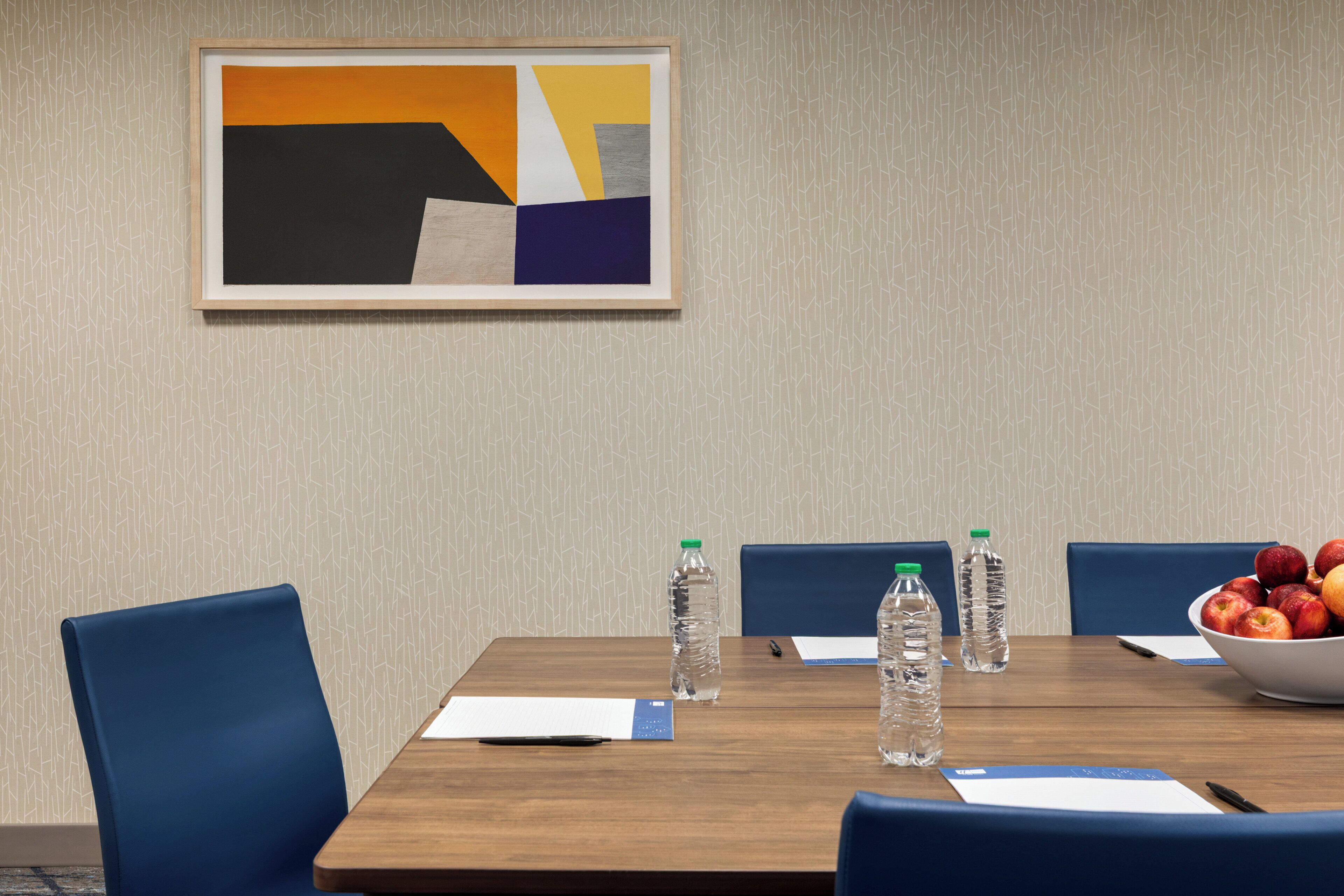 Une salle de conférence avec une table en bois, des chaises bleues, un tableau abstrait encadré, des bouteilles d’eau et un saladier rempli de fruits.