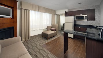 A compact hotel room with a kitchenette, a sofa, a television, and a large window with curtains.