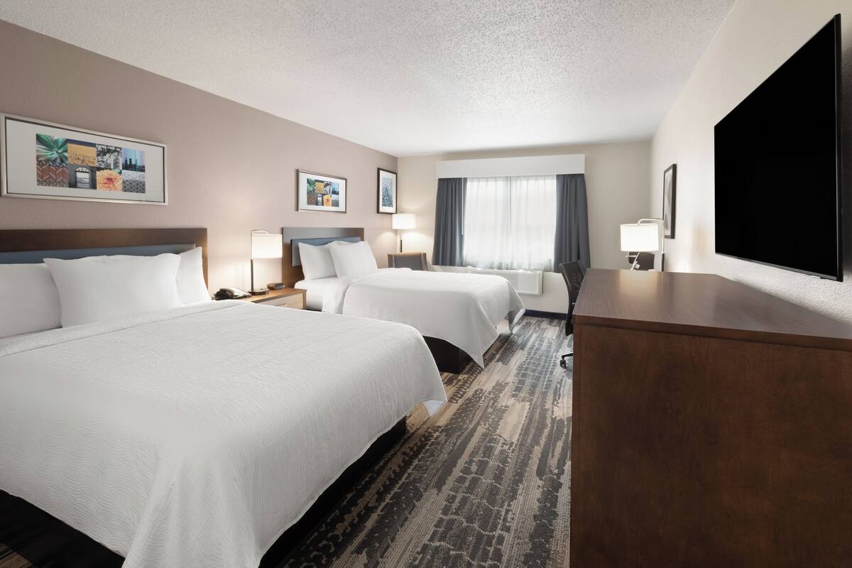 A hotel room with two beds, a flat-screen TV, a desk, and a large window with curtains.