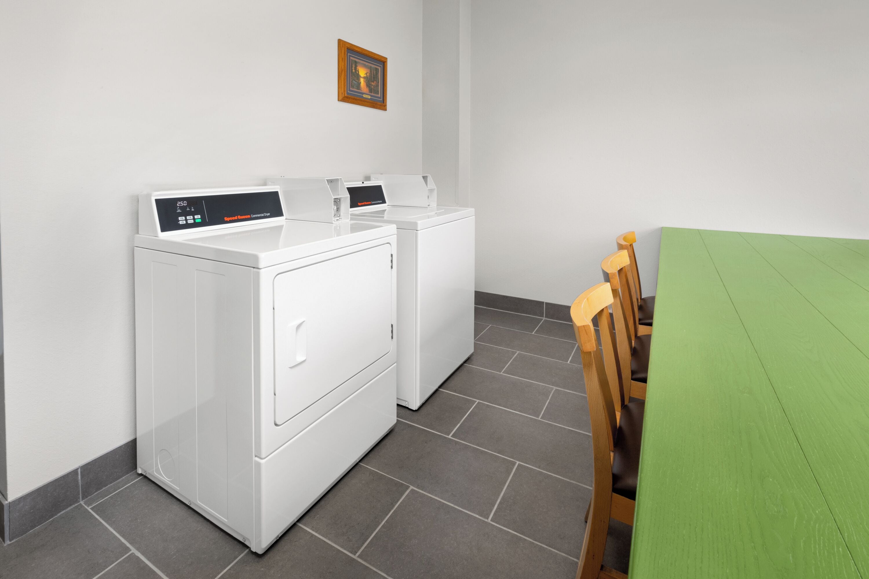 Een wasserette met twee witte wasmachines en een groene tafel met houten stoelen.