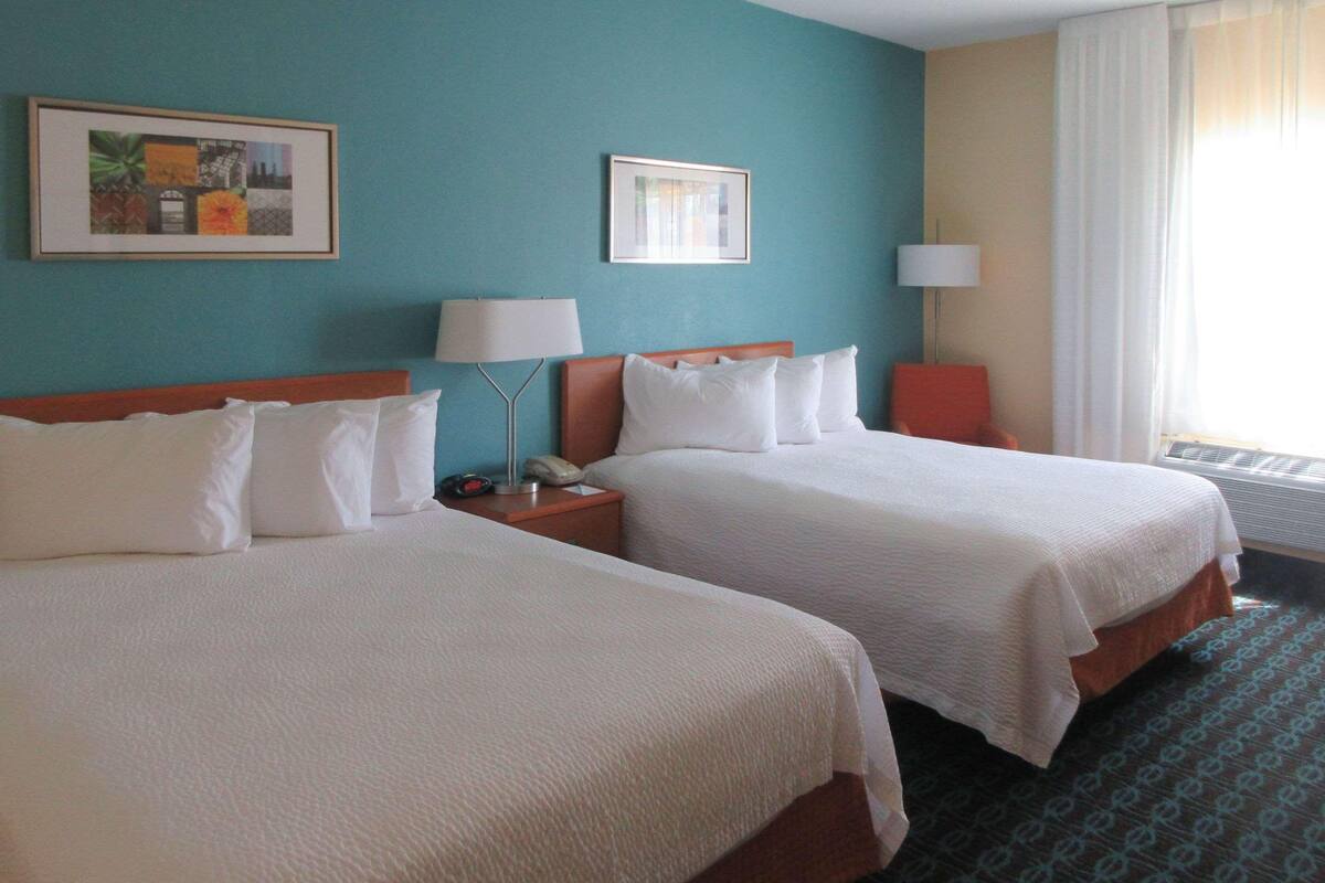 A hotel room with two beds, a nightstand with a lamp, a framed picture on the wall, and a window with curtains.