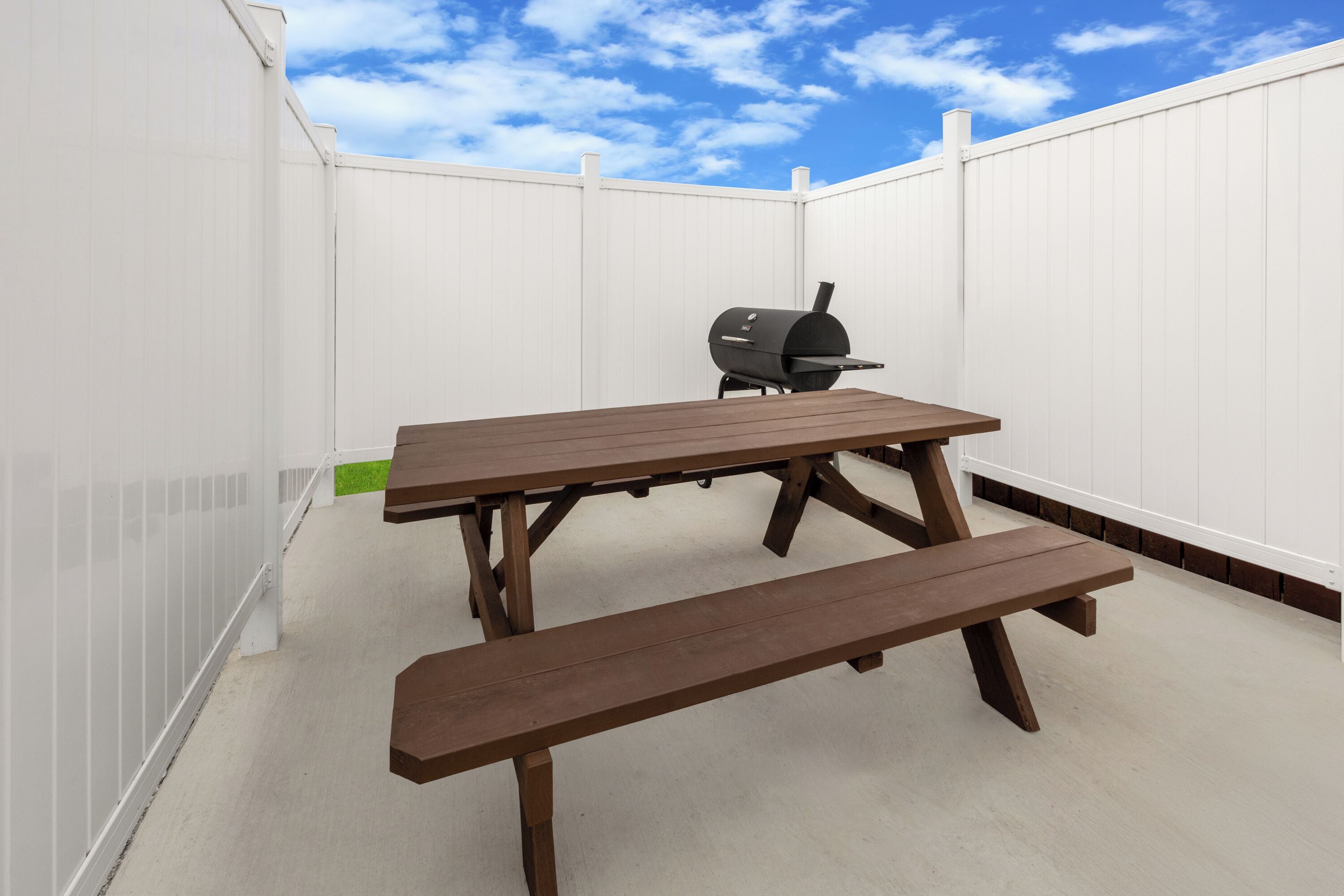 An outdoor patio with a wooden table and bench, a barbecue grill, and a white fence.