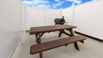 An outdoor patio with a wooden table and bench, a barbecue grill, and a white fence.
