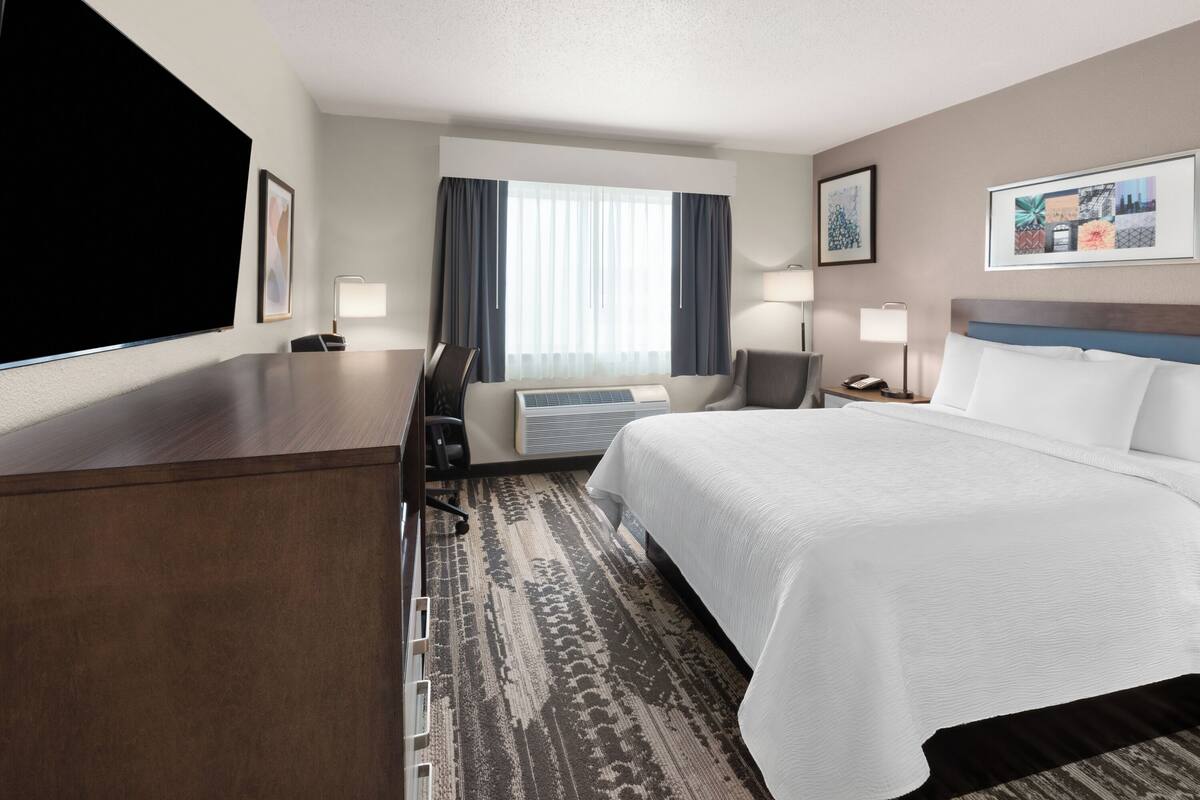 A hotel room with a large bed, a desk, a chair, a flat-screen TV, a window with curtains, and a wall-mounted air conditioner.