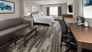 A hotel room with a gray sofa, a wooden desk, a bed with white linens, a large window with curtains, and artwork on the walls. - Wingate by Wyndham Marion (Marion)