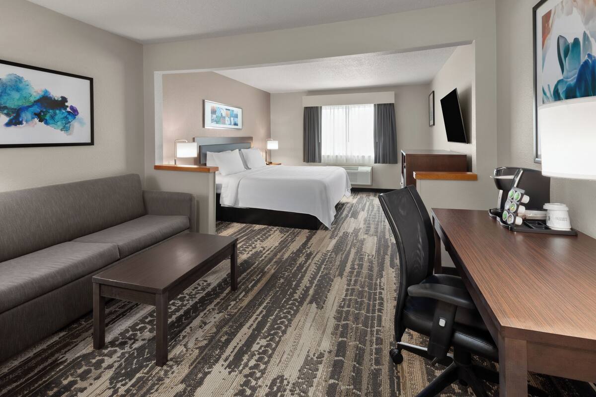 A hotel room with a gray sofa, a wooden desk, a bed with white linens, a large window with curtains, and artwork on the walls.