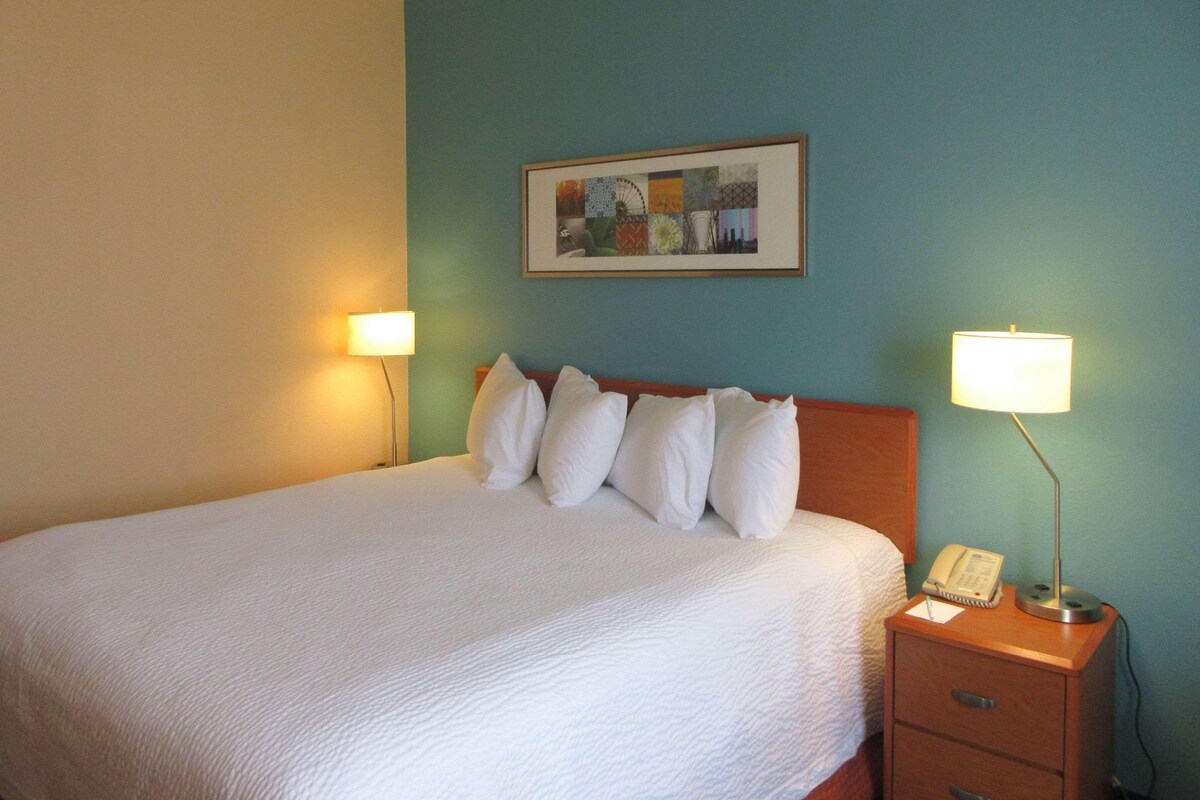 A neatly made bed with white bedding, a headboard, and a bedside table with a lamp and a phone.