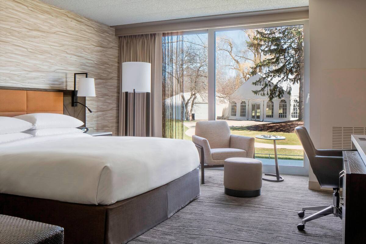 A hotel room with a large bed, a desk, a chair, and a view of a building and trees outside.