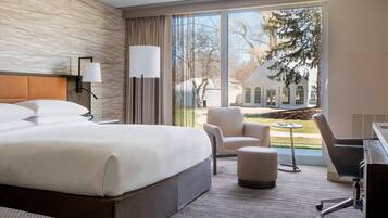 A hotel room with a large bed, a desk, a chair, and a view of a building and trees outside.