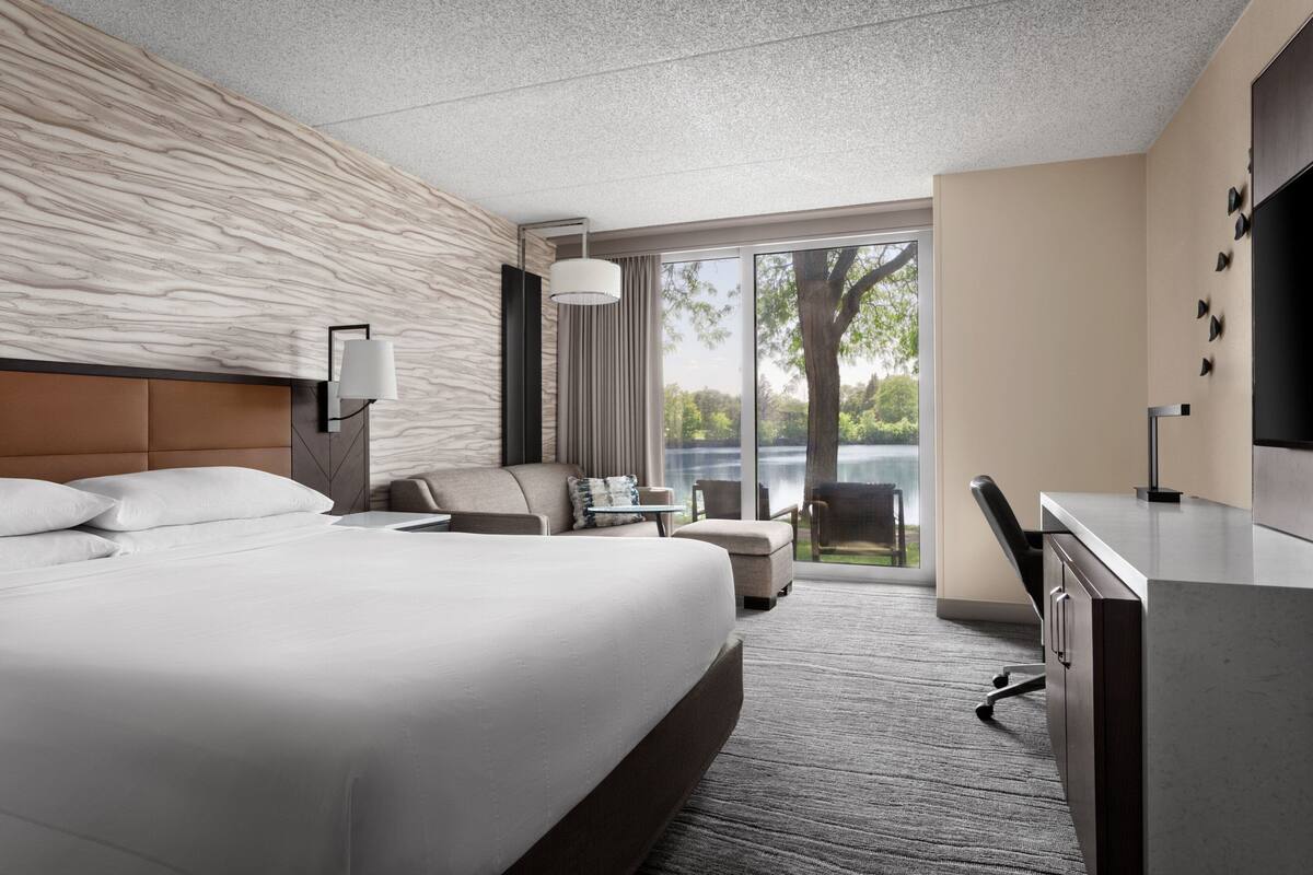 A modern hotel room with a large bed, a desk, a chair, and a view of the outdoors.
