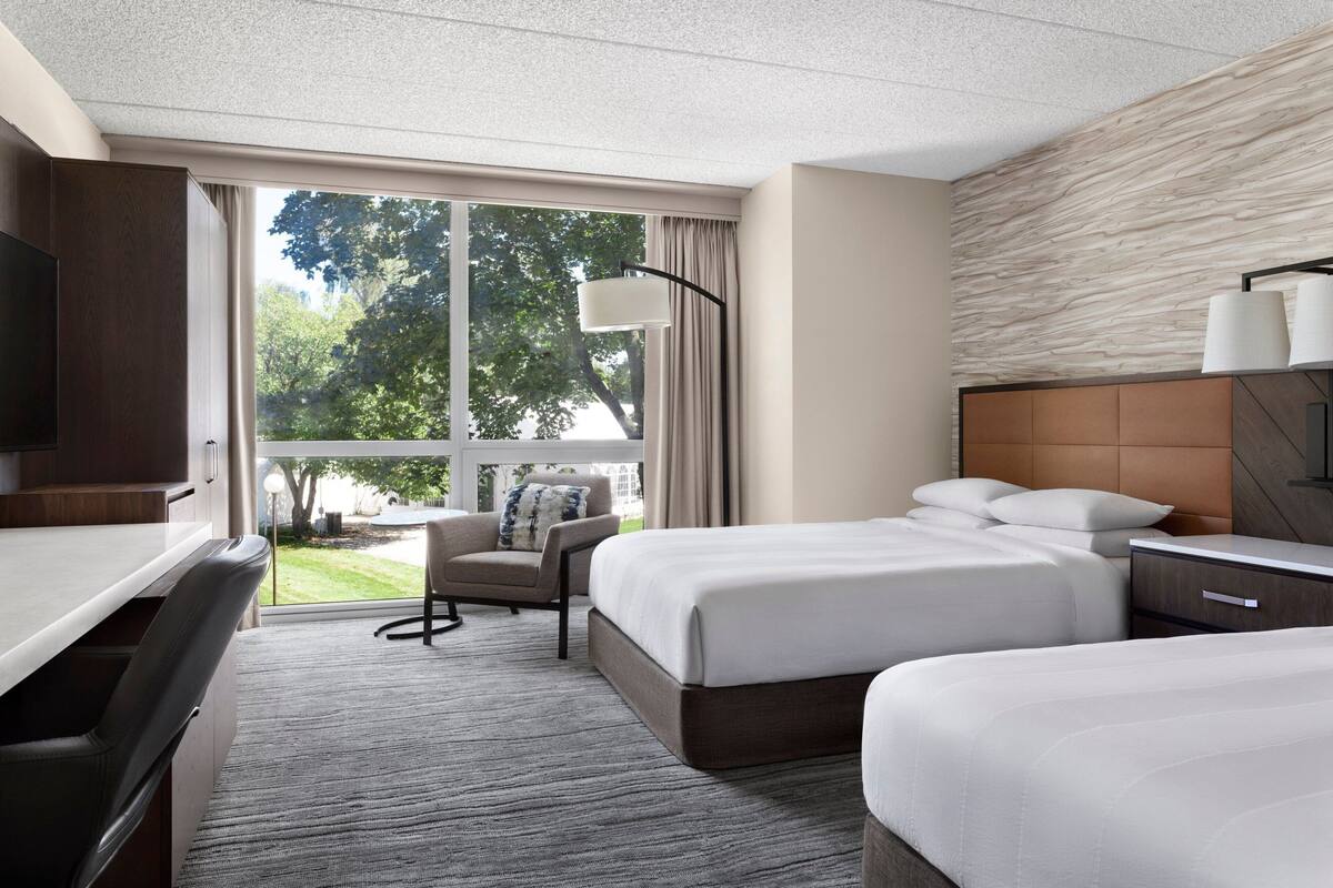 A hotel room with two beds, a desk, a chair, a TV, and a large window with a view of trees.