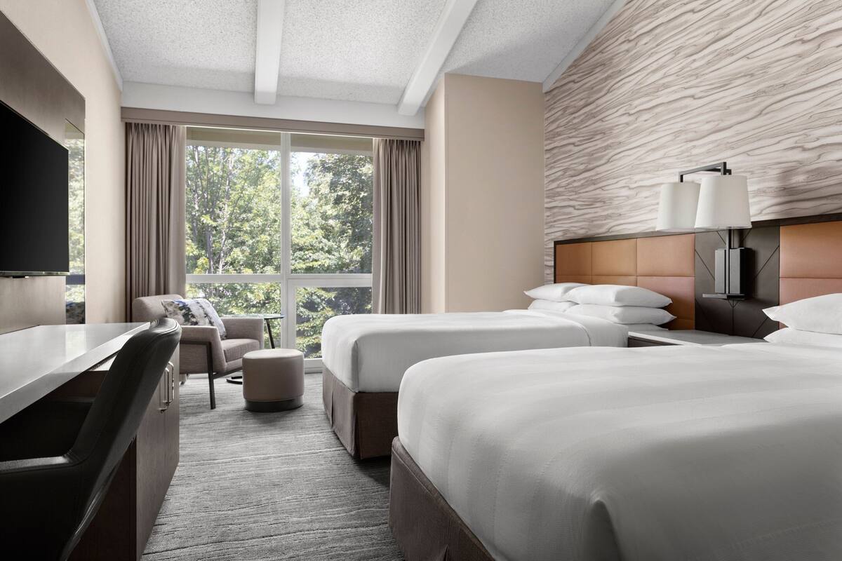 A hotel room with two beds, a desk, a TV, a sofa, and a window with a view of trees.