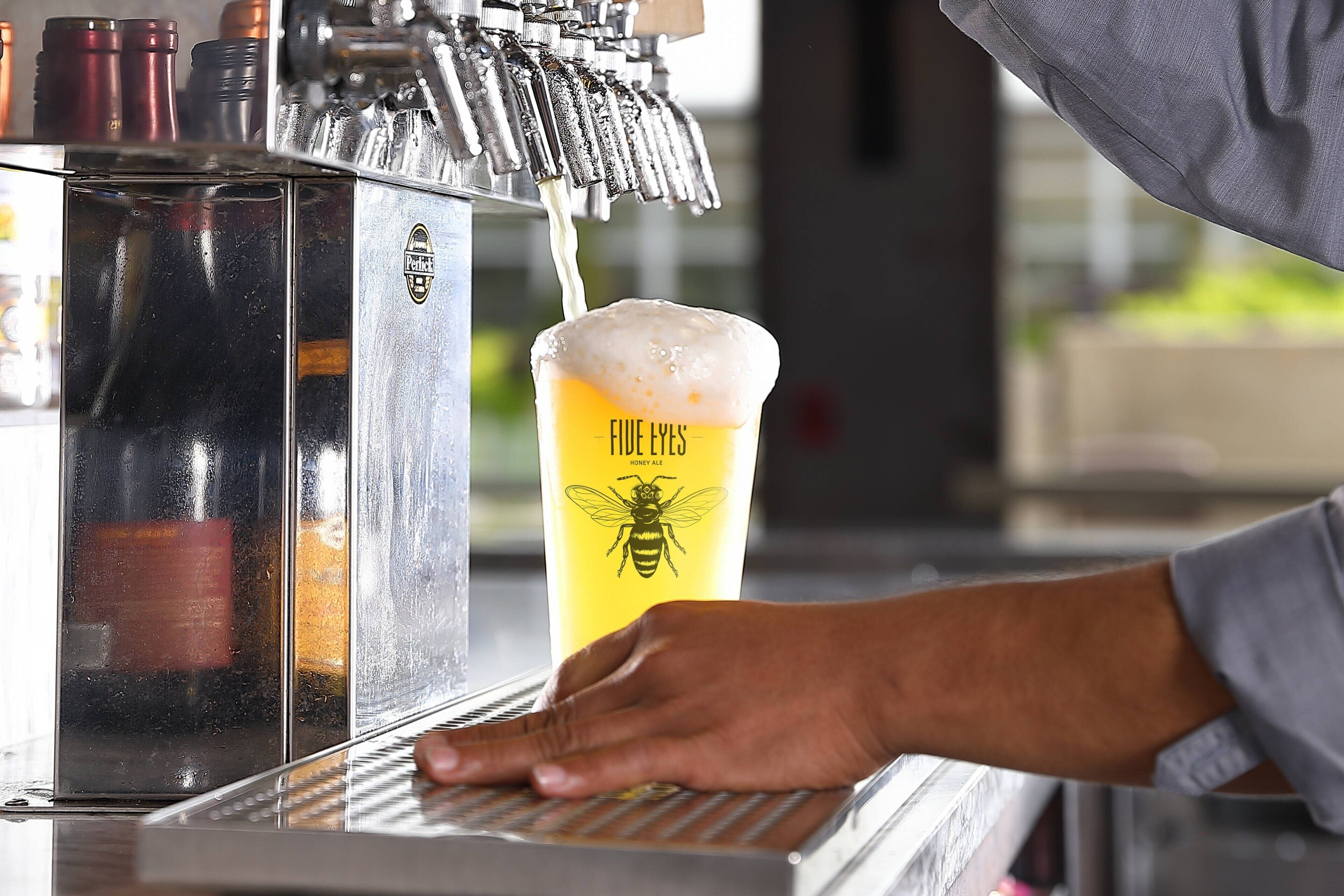 A hand operating a beer tap, pouring a beer into a glass labeled "FIVE EYES Honey Ale."