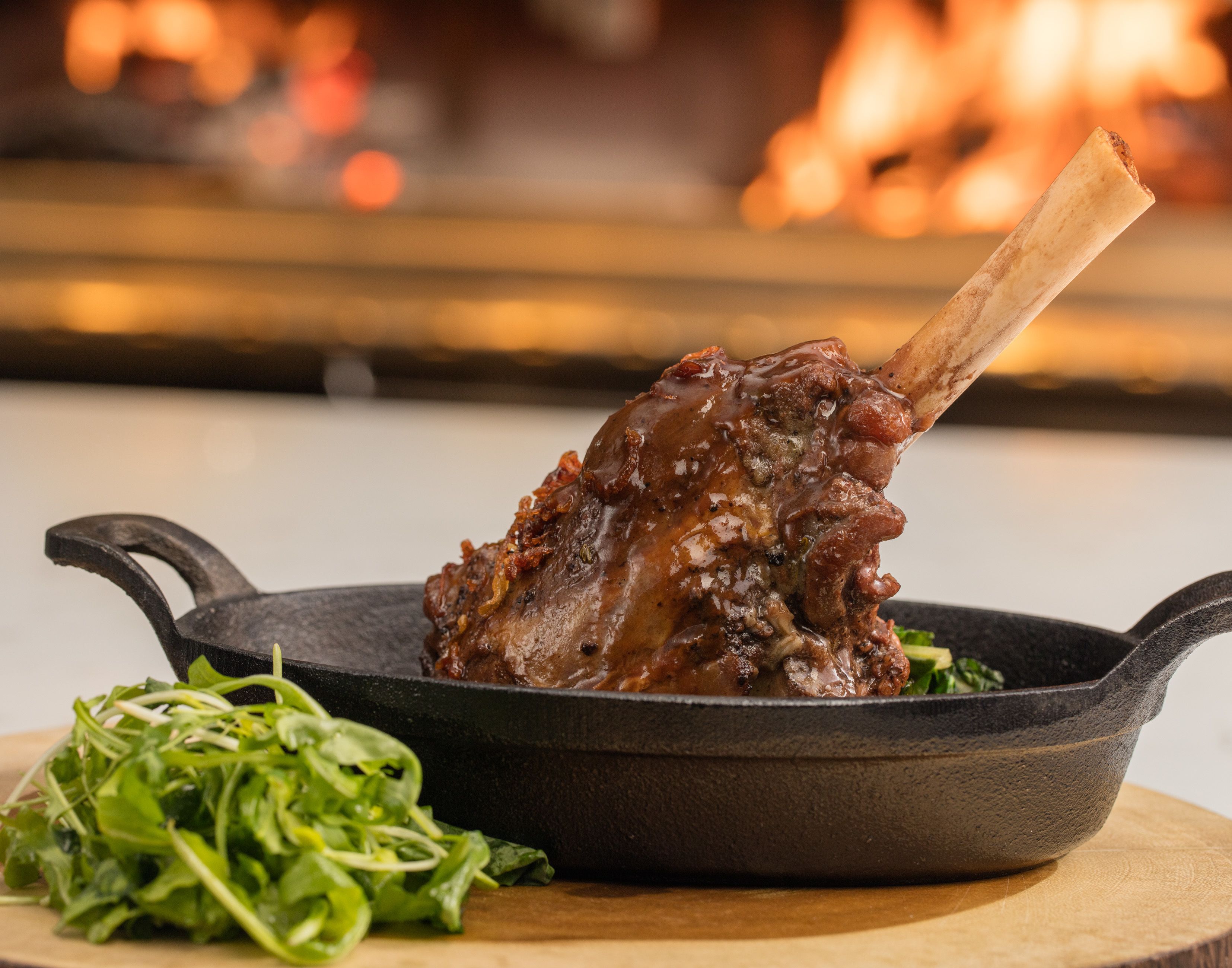 Una sartén de hierro fundido con un palillo de cordero asado y verduras sobre una tabla de madera.