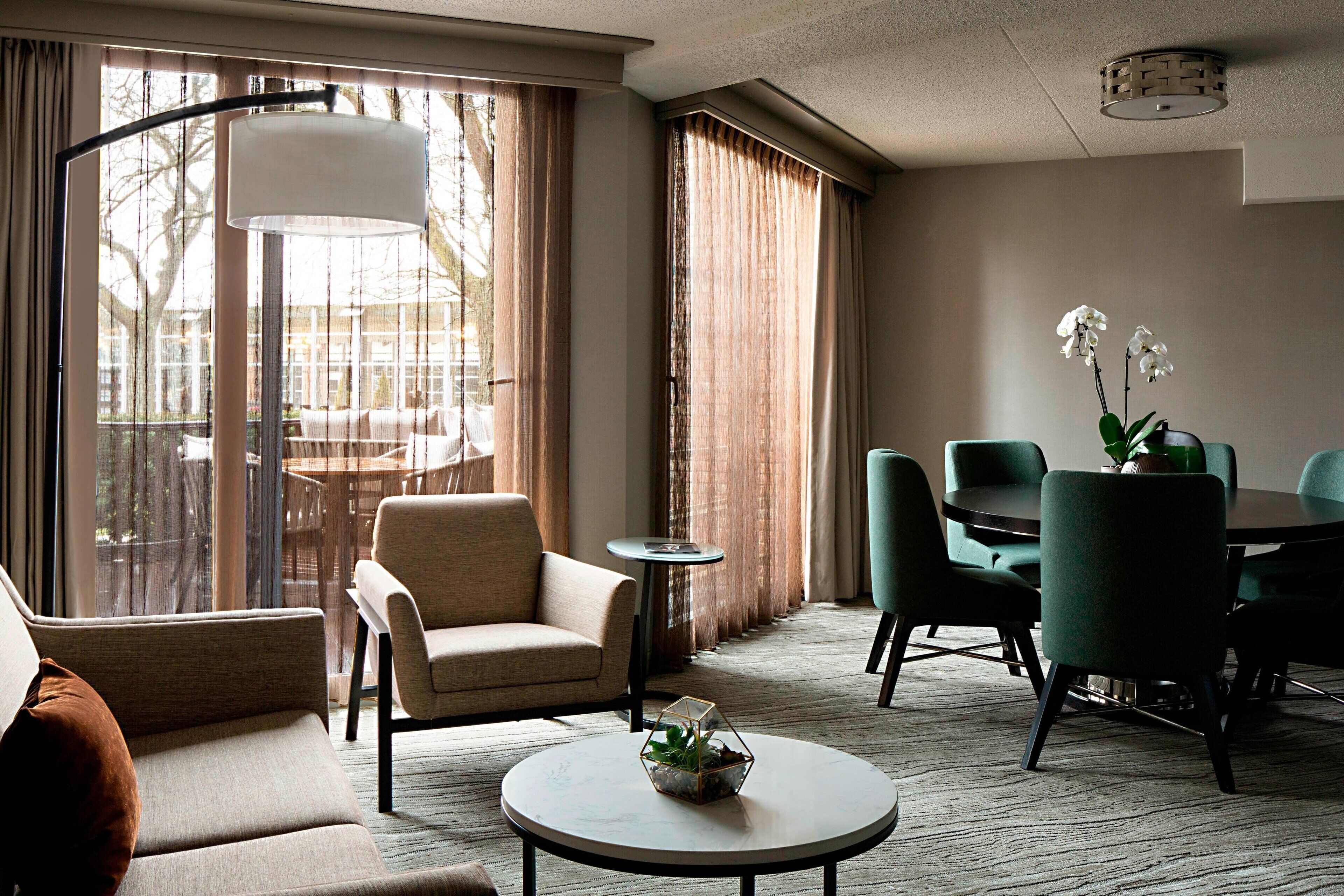 A hotel room with a sofa, a round coffee table with a plant, a dining table with chairs, and a large window with curtains.