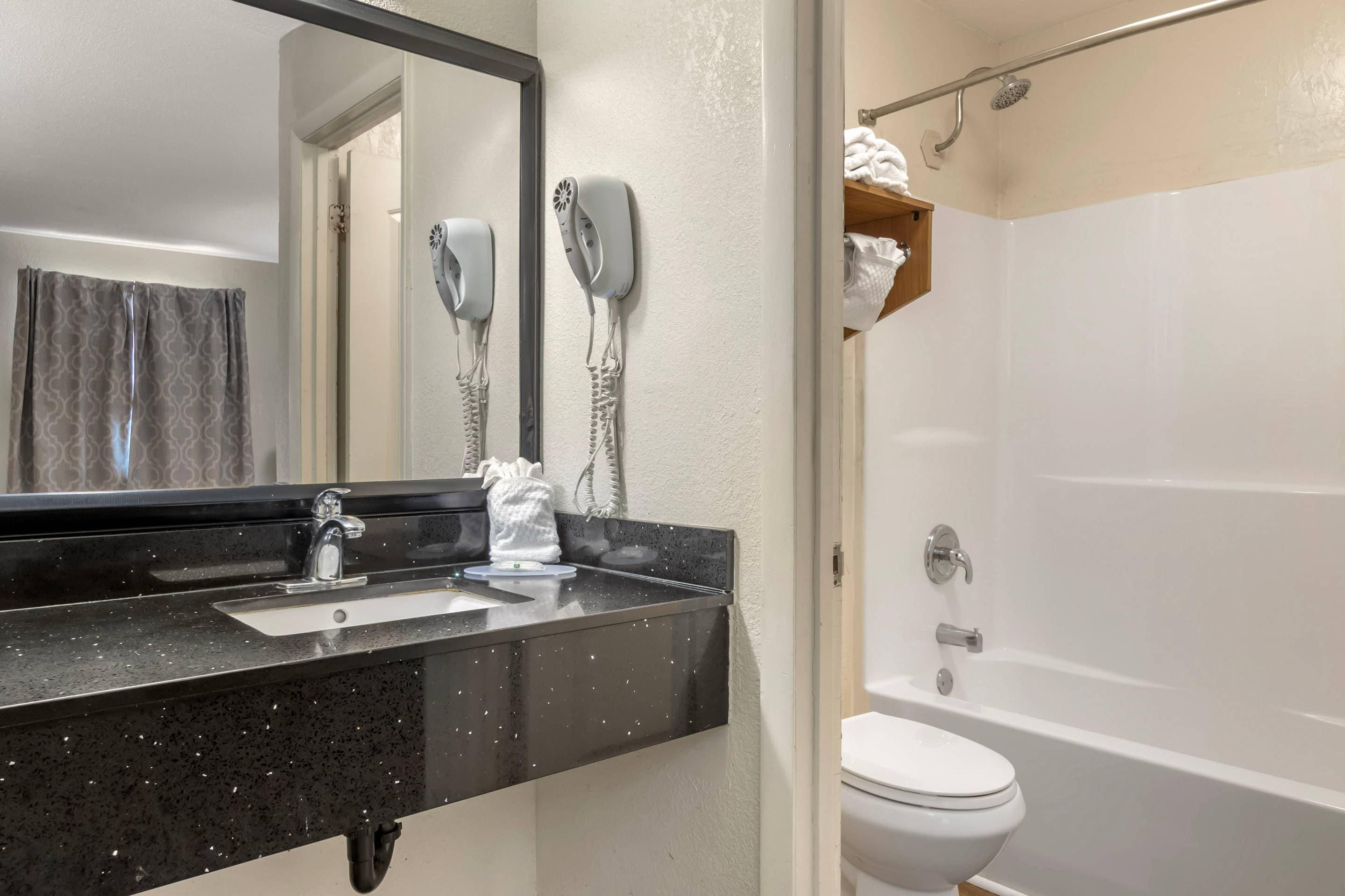 A bathroom with a black countertop, a white sink, a mirror, two hair dryers mounted on the wall, a white toilet, and a bathtub.