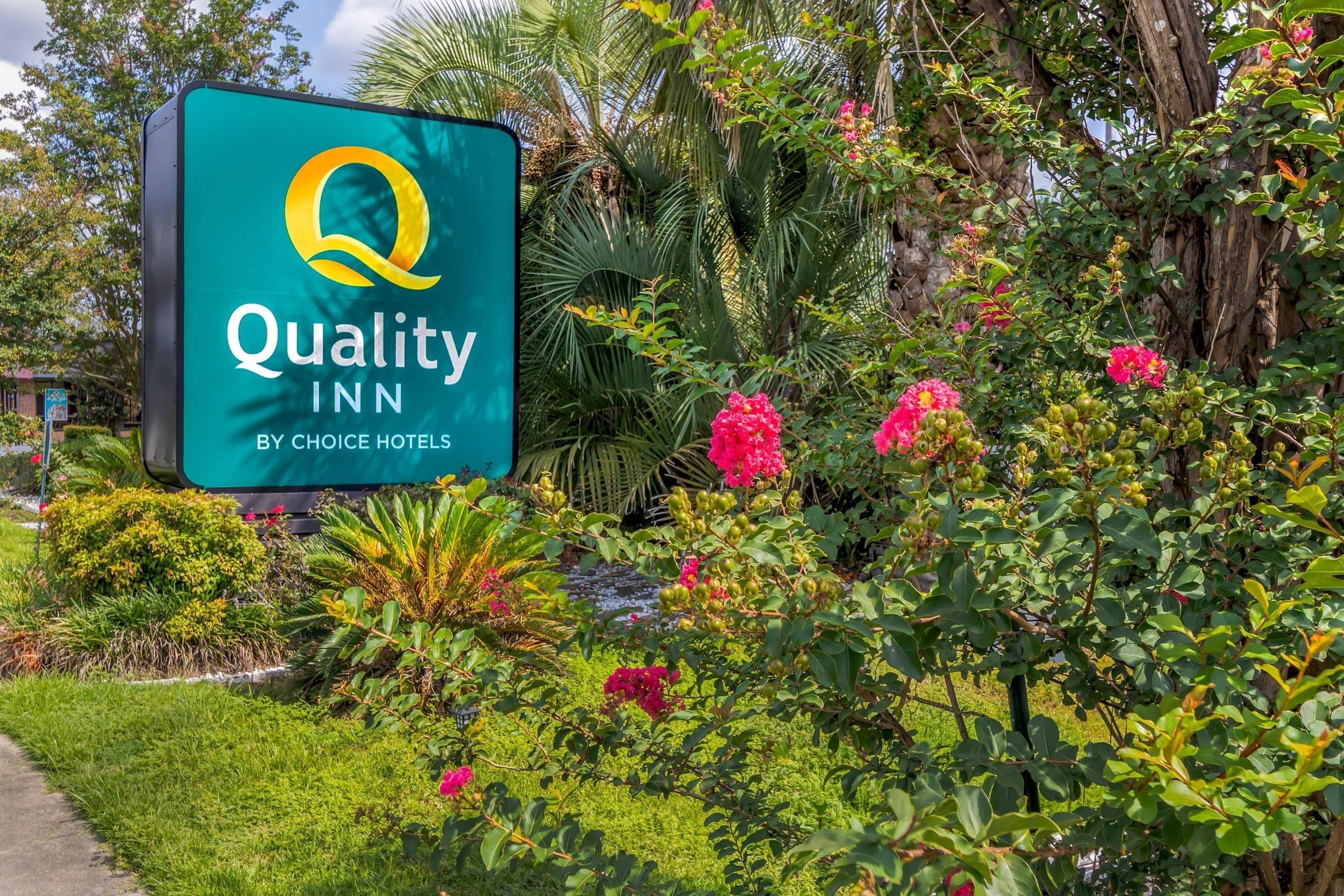 A sign for Quality Inn by Choice Hotels surrounded by lush greenery and flowering plants.