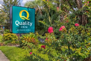 Placa da Quality Inn by Choice Hotels em meio a vegetação exuberante e plantas floridas.
