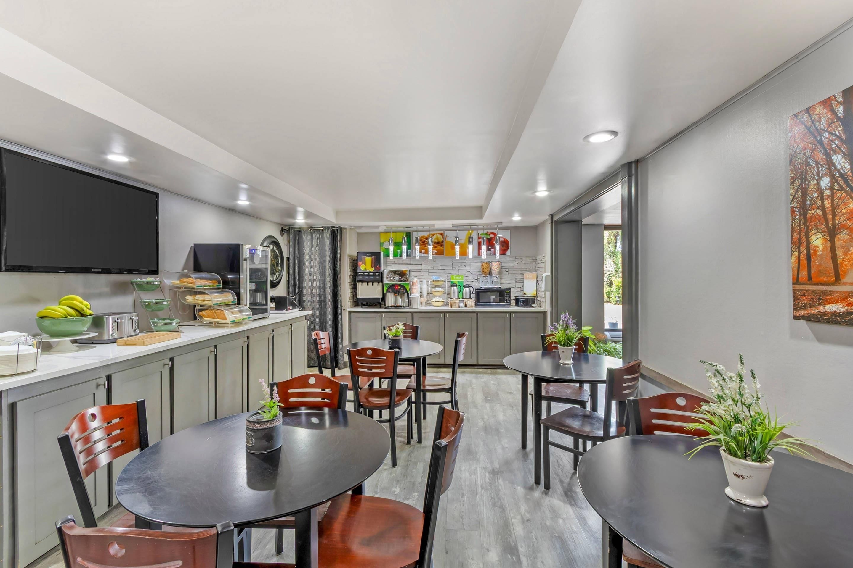 Una cocina moderna con televisor de pantalla plana, refrigerador y microondas. Hay dos mesas de comedor con sillas, y una ventana con vista a árboles.